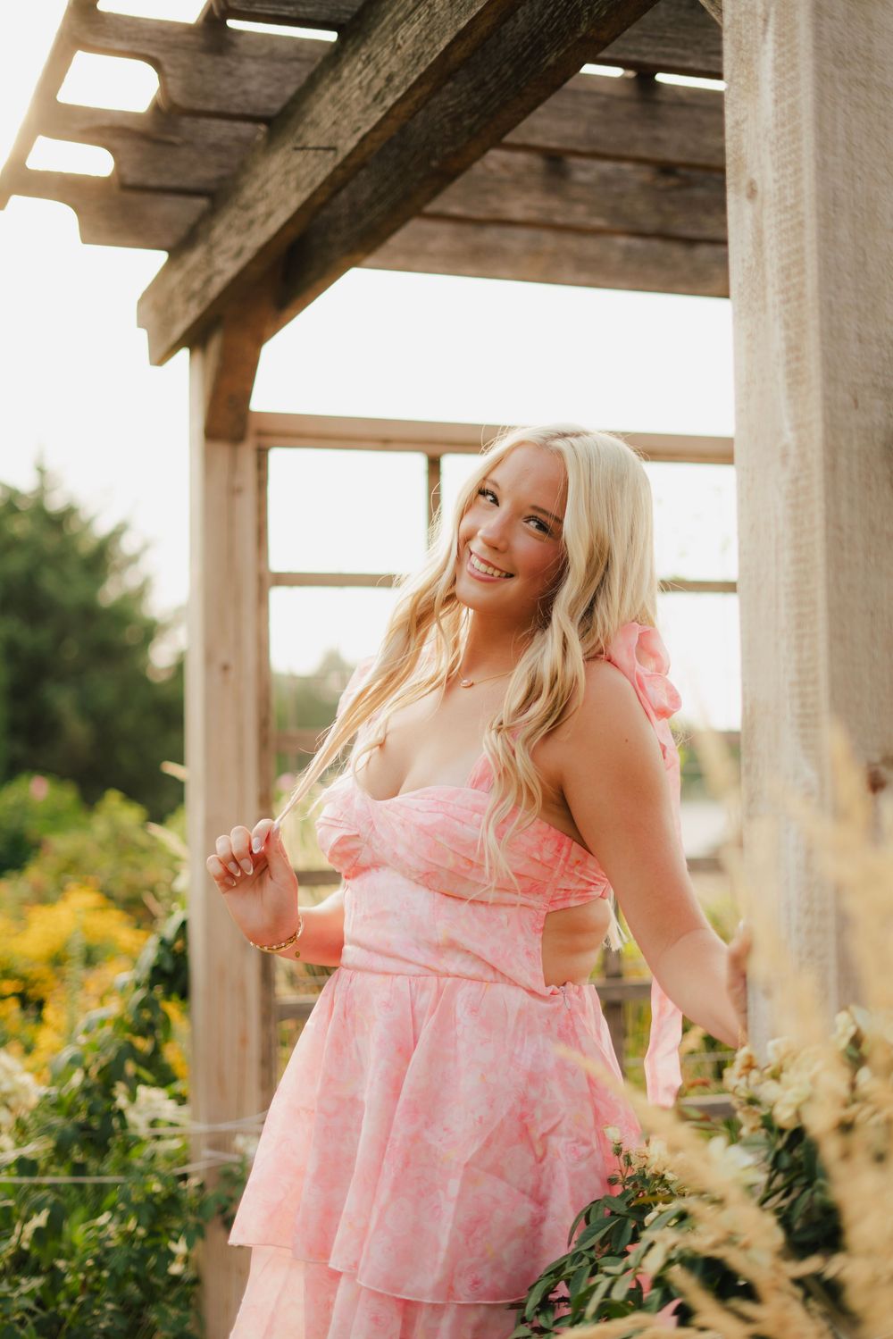 Smiling blonde woman in a pink floral dress posing under a wooden pergola surrounded by garden flowers.