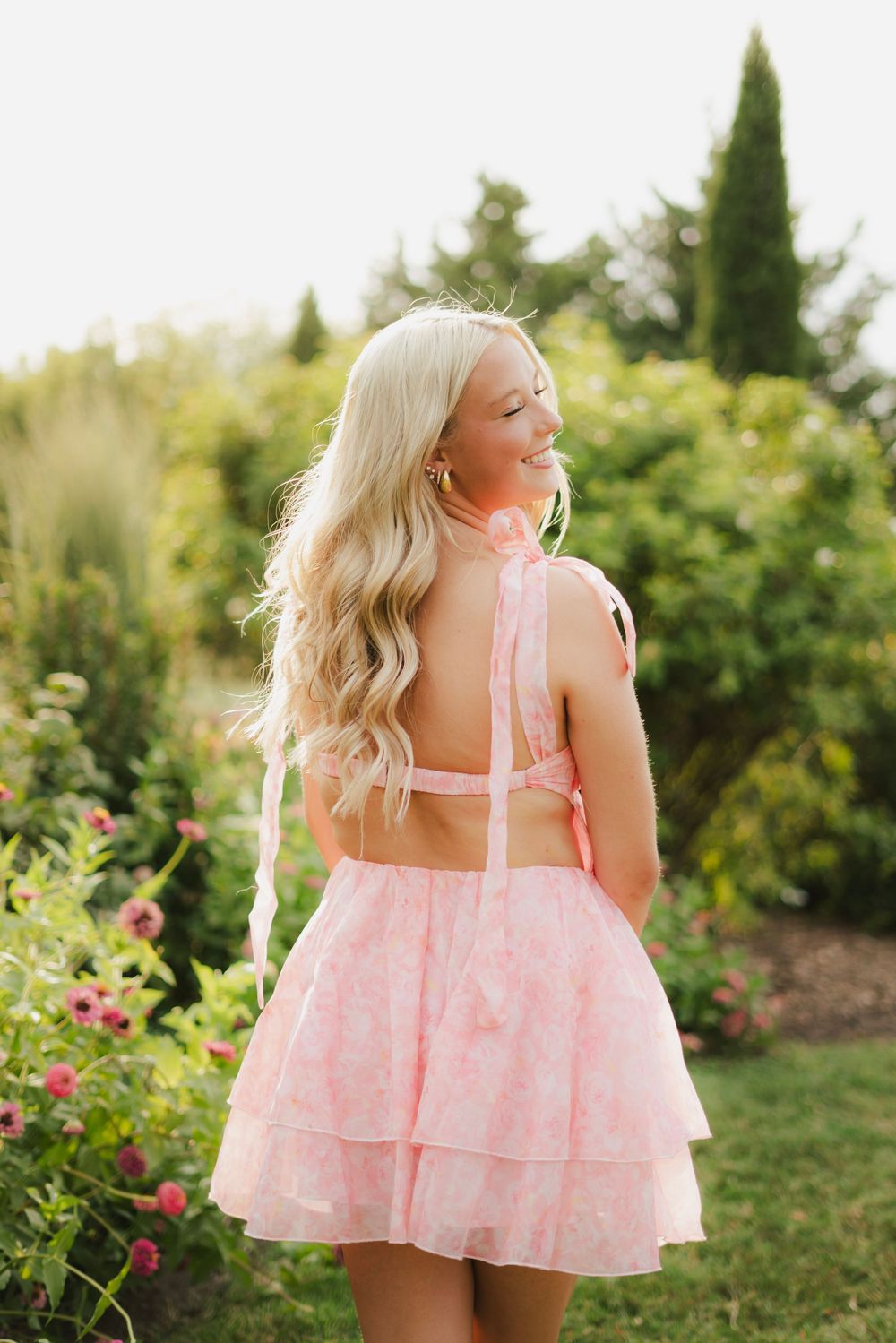 Blonde woman in pink tiered backless dress standing in lush garden with flowers and greenery.