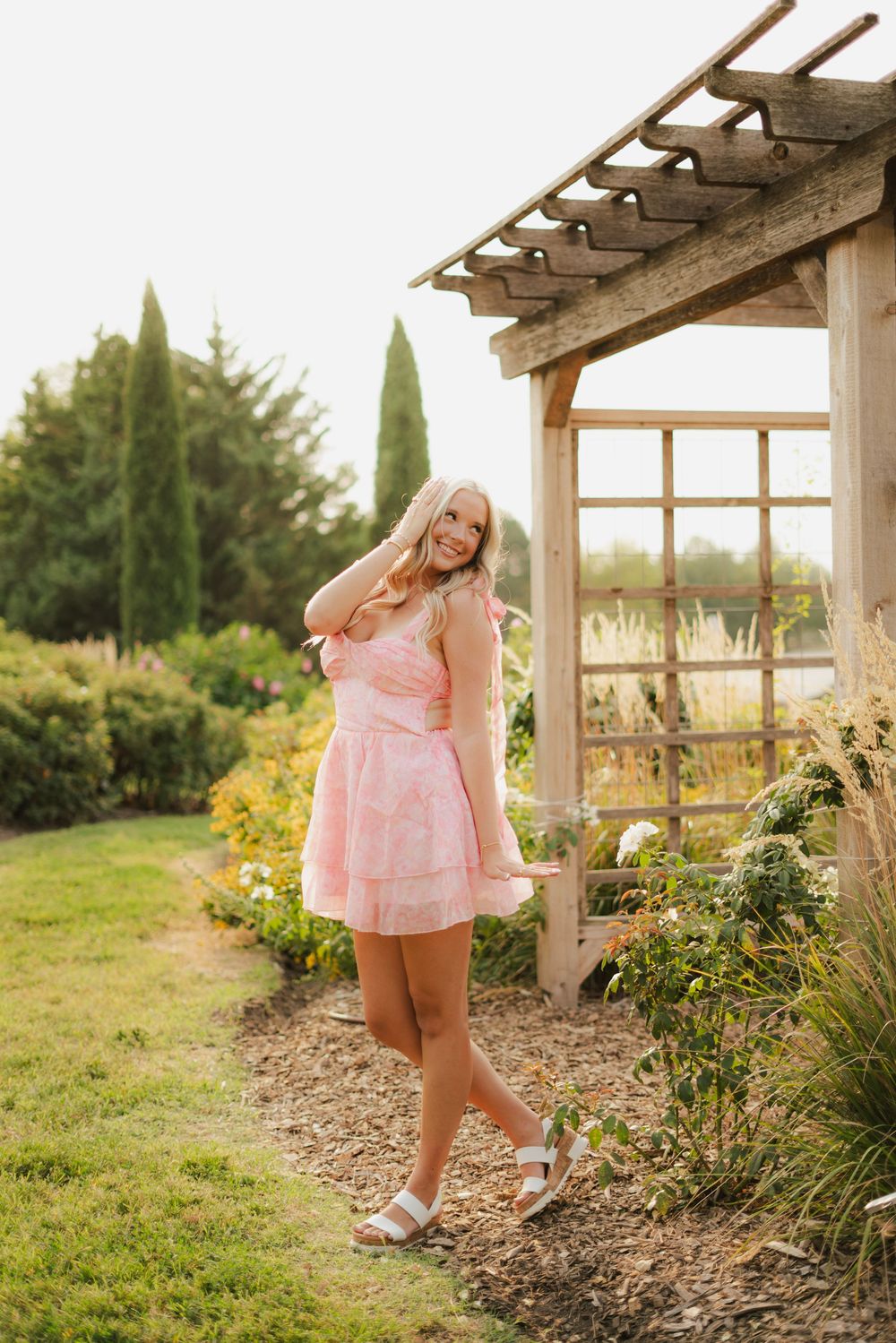 Blonde woman in pink mini dress and white sandals posing in a garden near a wooden pergola.
