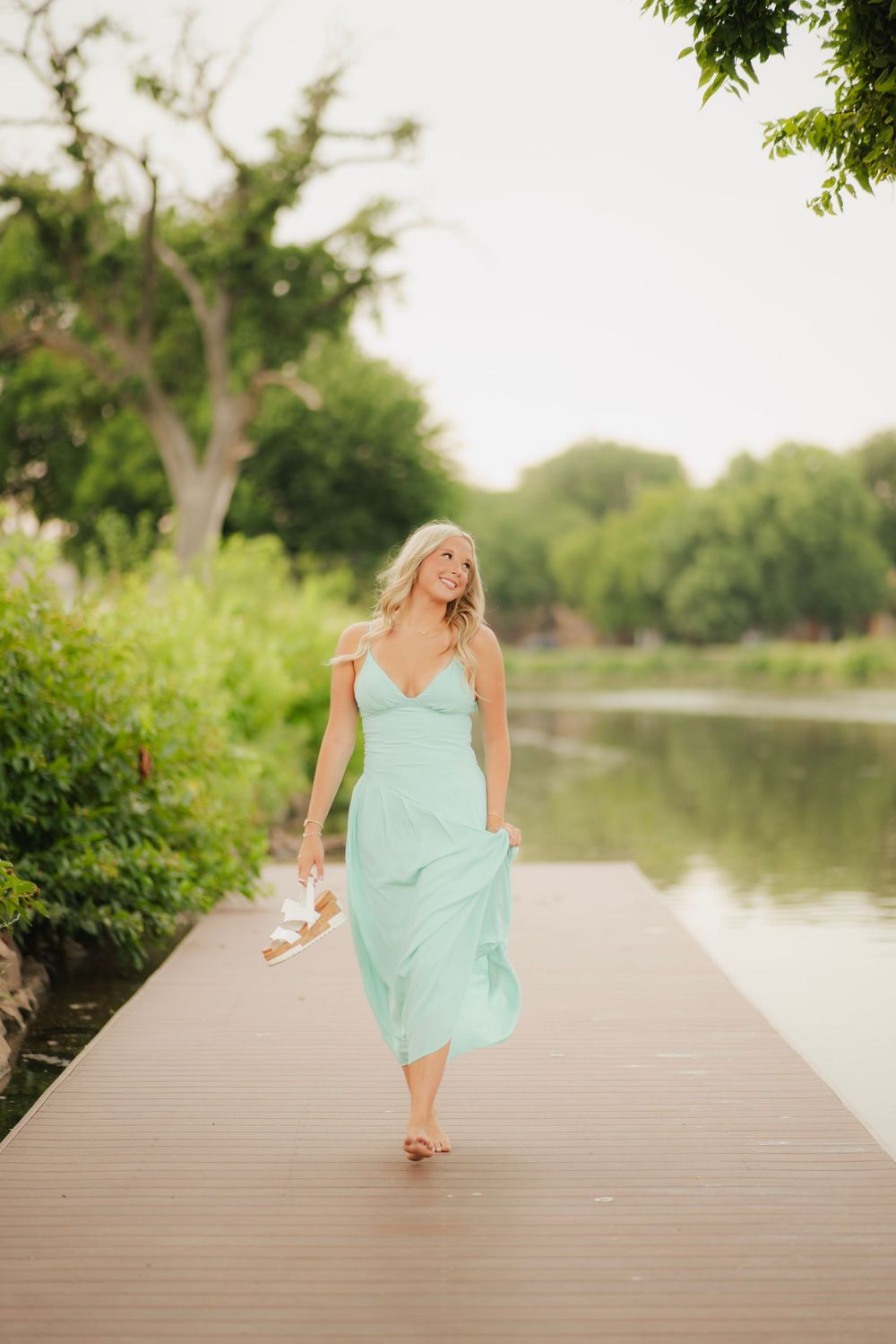 Blonde woman in mint green midi dress walking barefoot on wooden dock by serene lake surrounded by lush greenery.