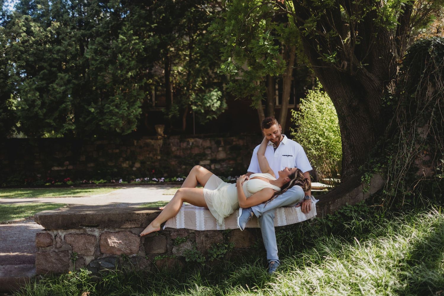 Couple relaxing outdoors on stone bench under large tree, woman reclining on man's lap in garden setting.