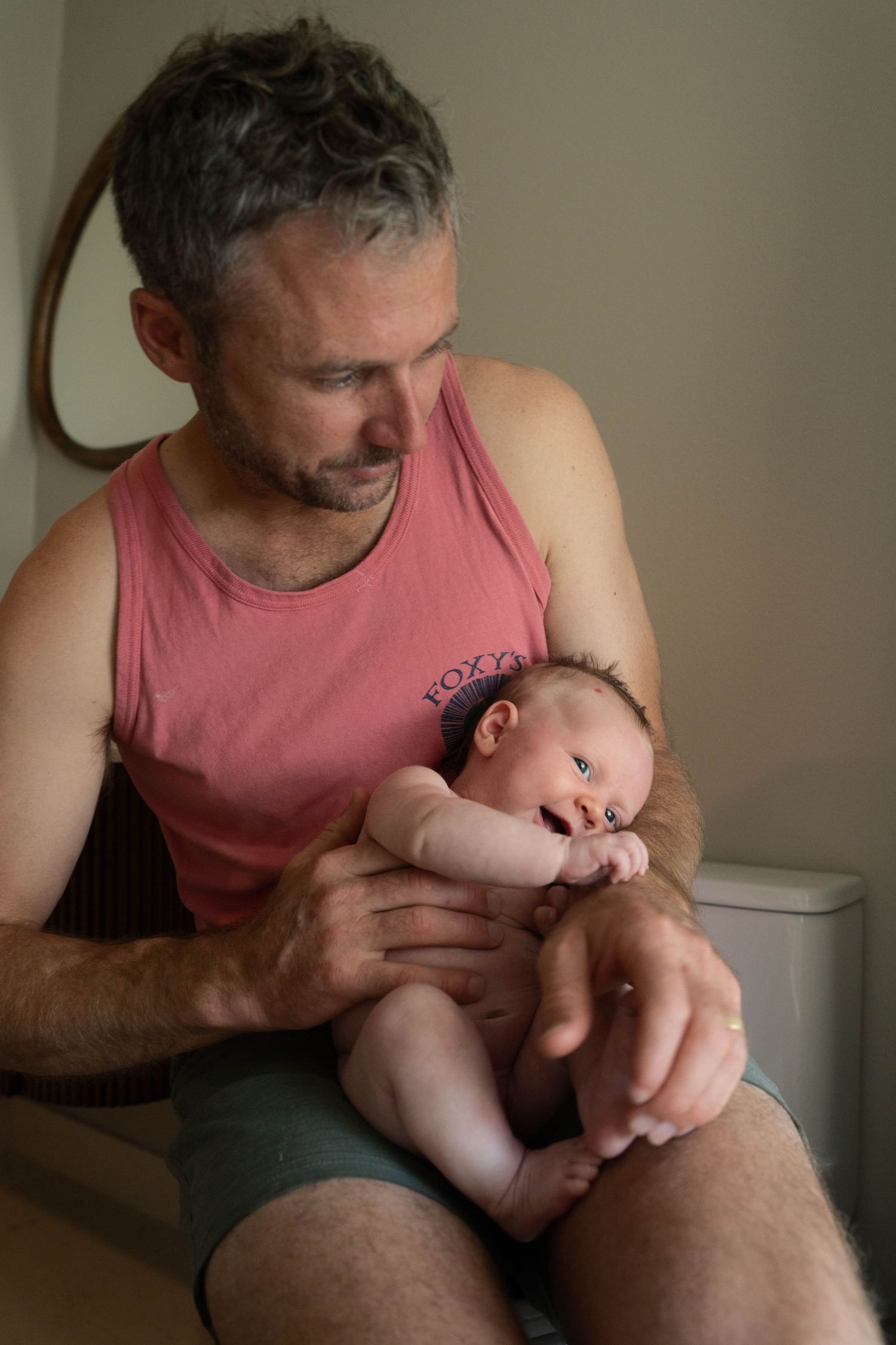 A middle-aged father in a pink tank top tenderly holds his newborn baby while seated indoors.