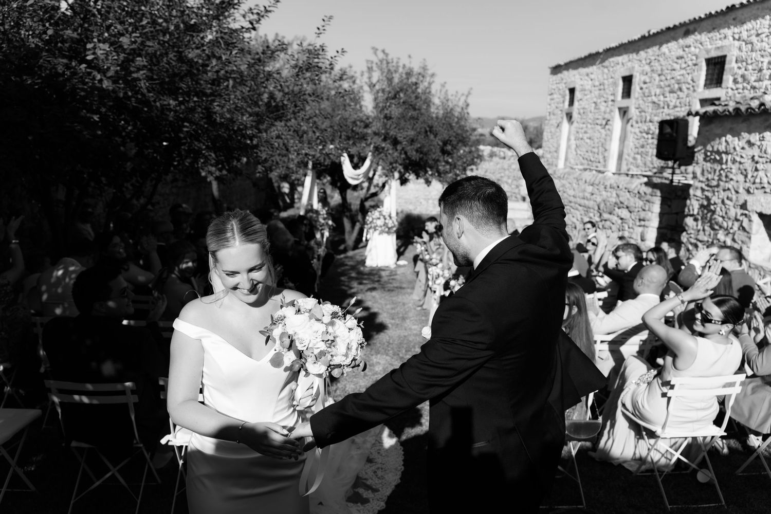 Black and white photo of a bride and groom walking past cheering guests at an outdoor wedding ceremony.