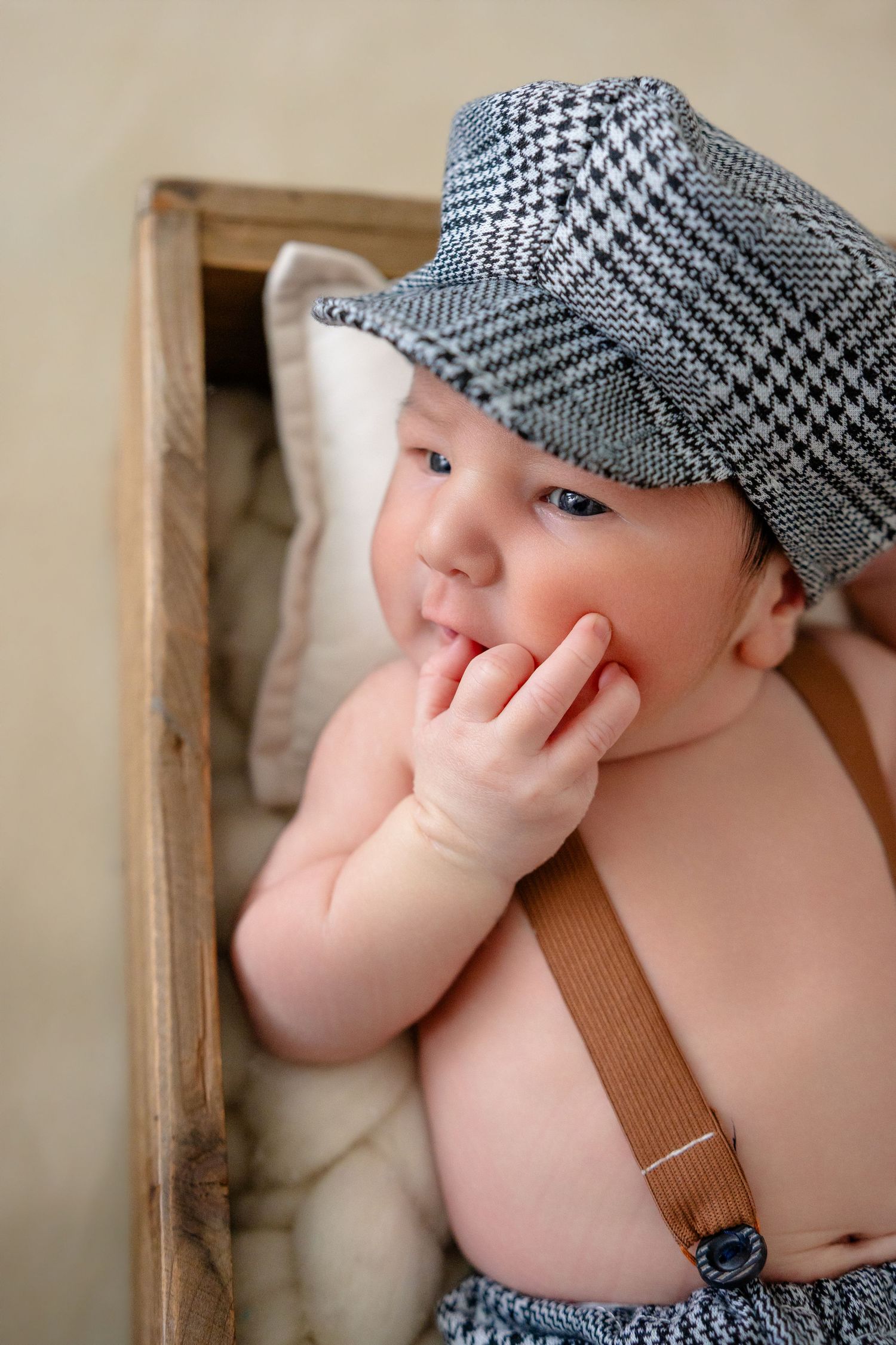 Adorable baby wearing a houndstooth newsboy cap and brown suspenders lying in a wooden box.