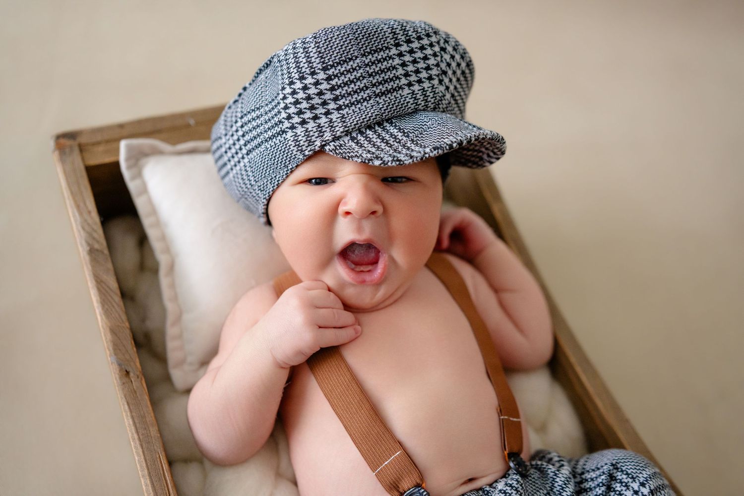 Adorable yawning baby wearing a plaid newsboy cap and suspenders lying in a rustic wooden box.