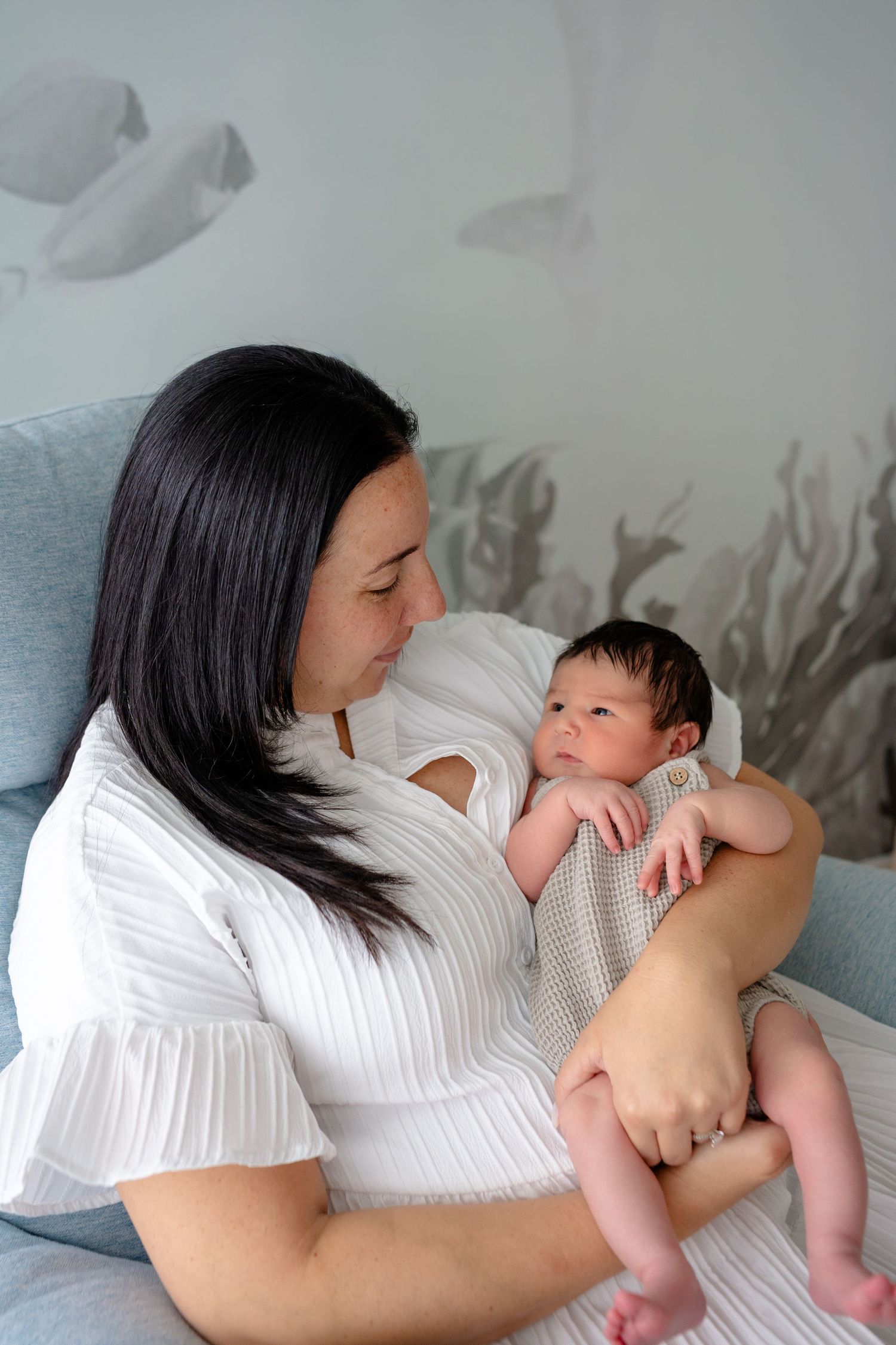 A mother in a white dress tenderly holds her newborn baby in a softly decorated nursery room.
