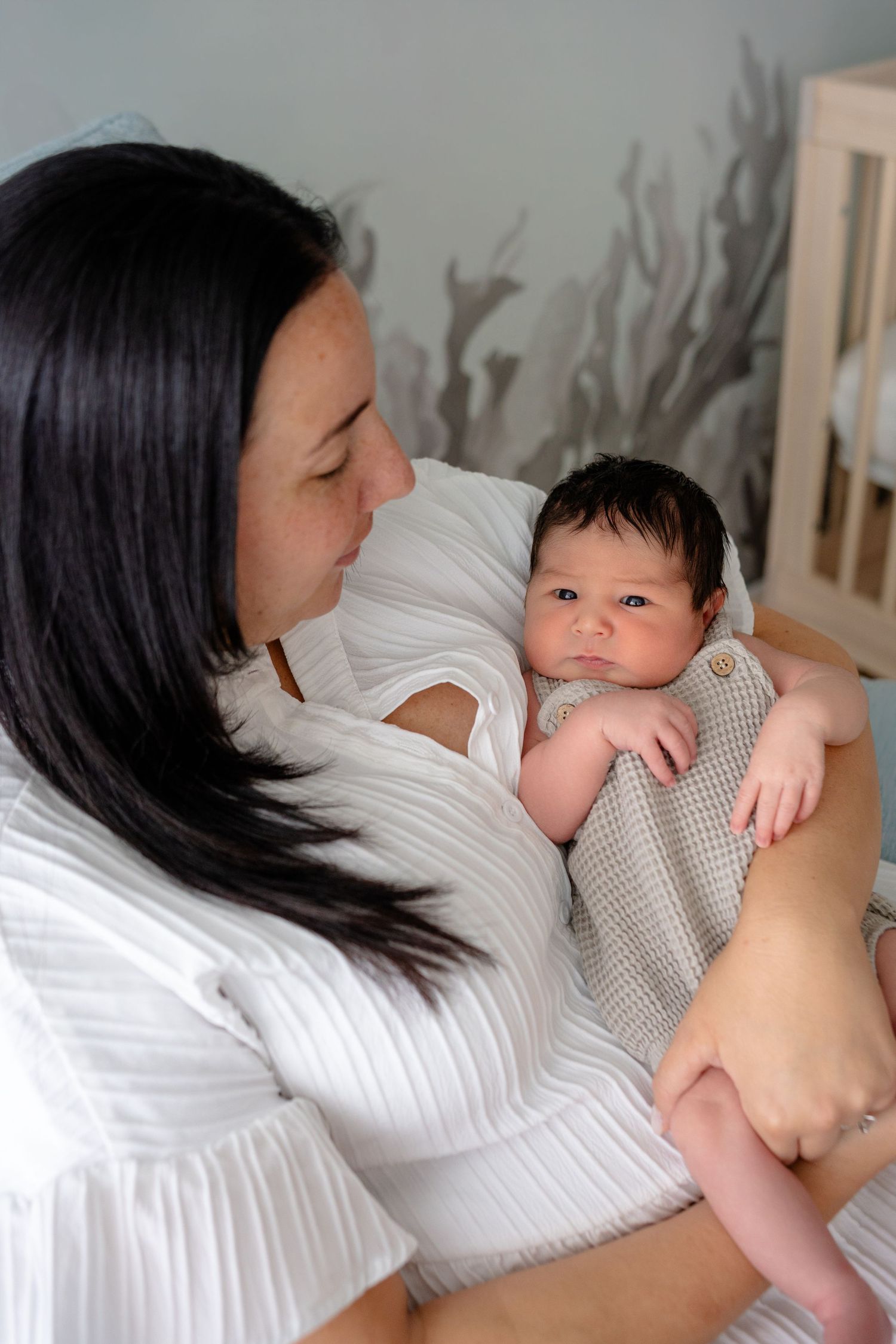 Mother in white dress lovingly gazes at her newborn baby in a softly decorated nursery room.