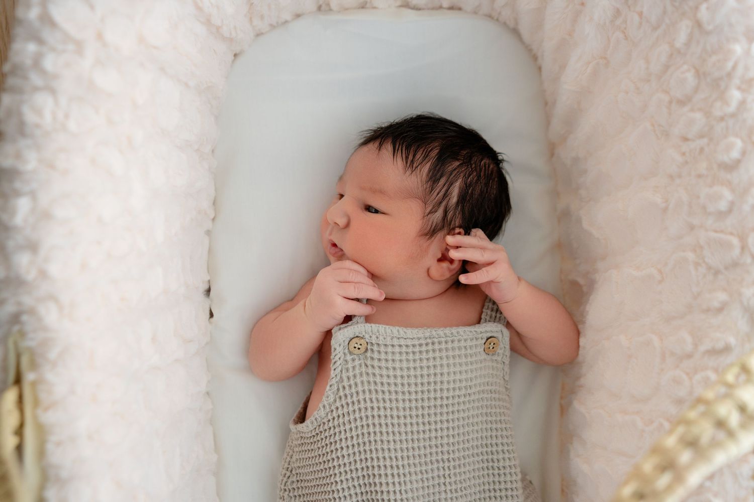 Newborn baby in sage green waffle romper lying peacefully in a white wicker bassinet.