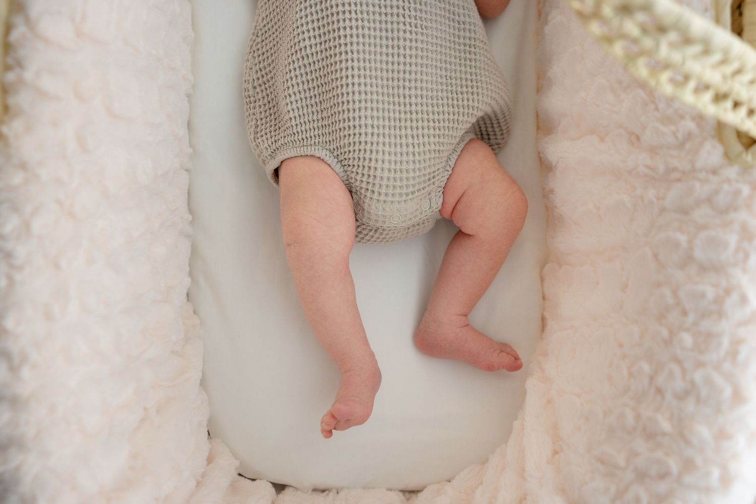 Close-up of a newborn baby's legs resting in a soft white bassinet, wearing a neutral knit romper.