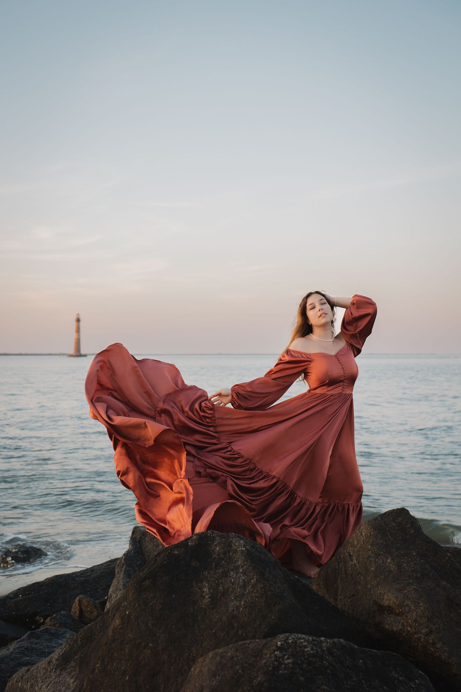 A woman in a flowing rust colored dress at sunset. She stands upon the rocks with the ocean and Morris Island lighthouse behind her.