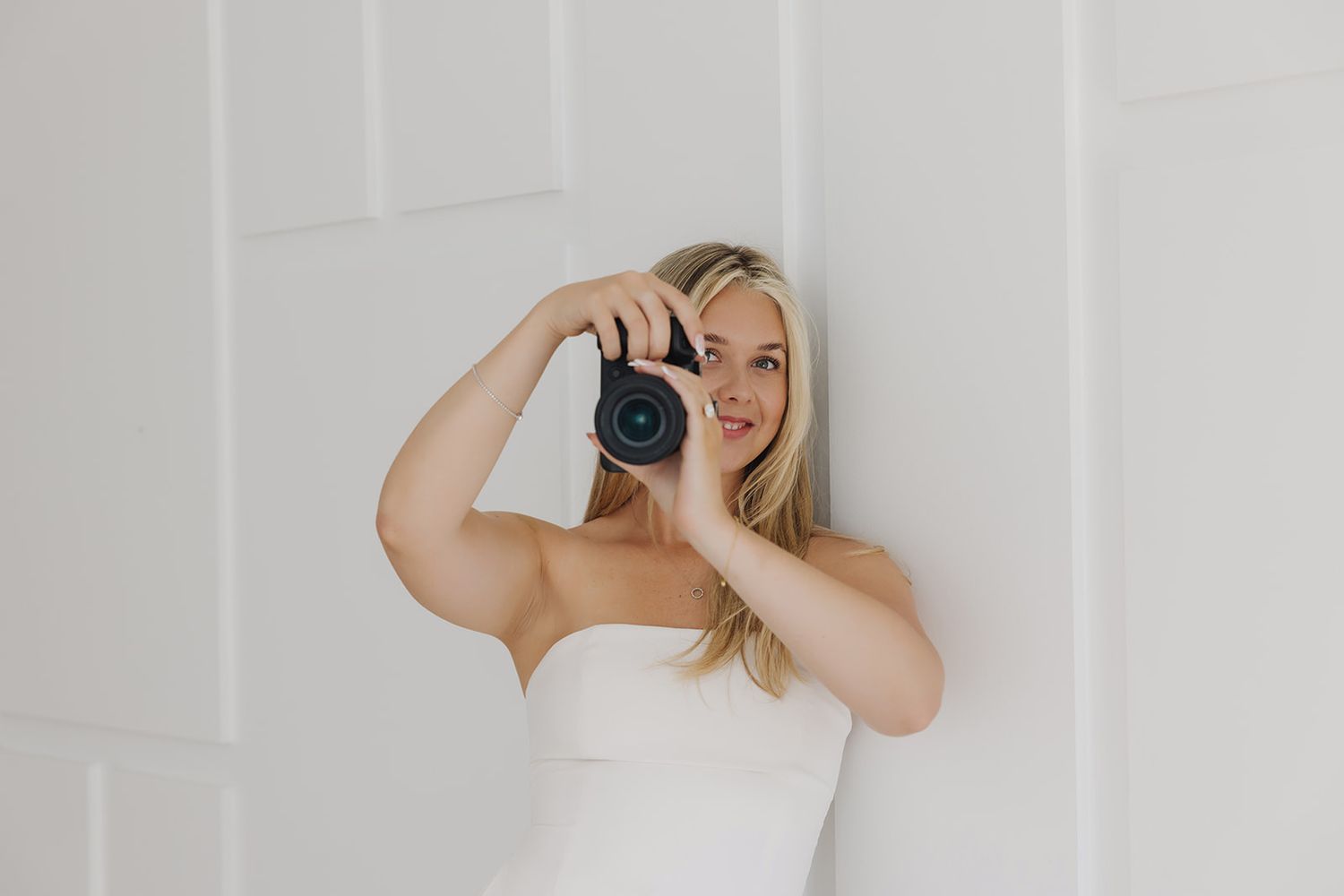 Blonde woman in white strapless dress holding a DSLR camera up to her eye against a white wall.
