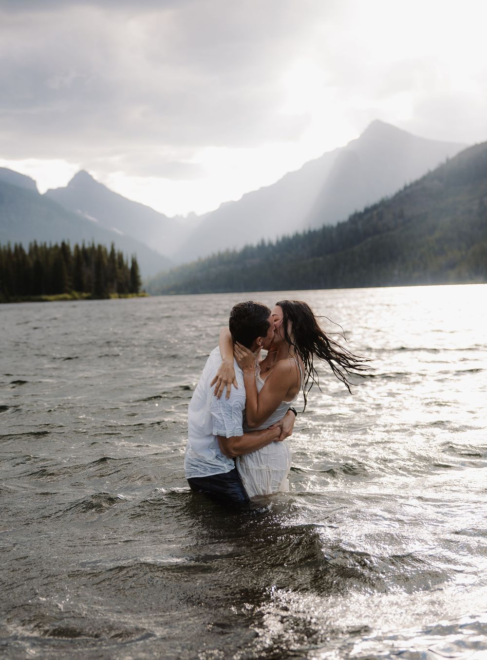 Couple kisses while standing in mountain lake with dramatic sunlit sky above.