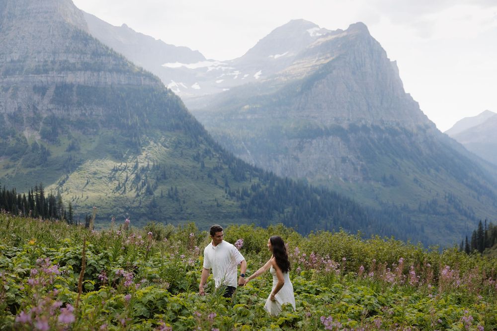 Couple holds hands walking through wildflowers with stunning mountain peaks in Glacier National Park.