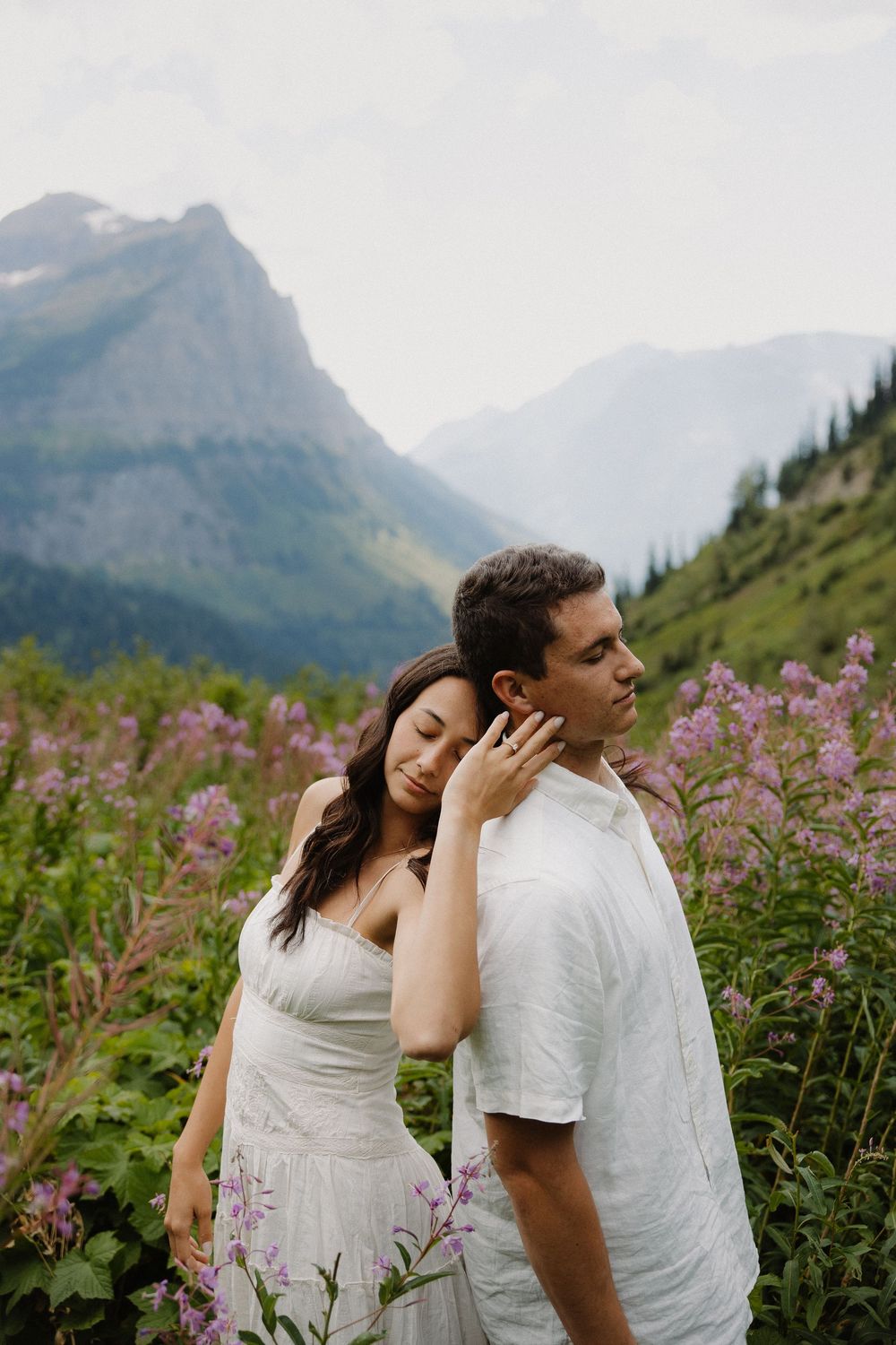 Romantic couple embraces in a field of pink wildflowers with dramatic mountain backdrop.