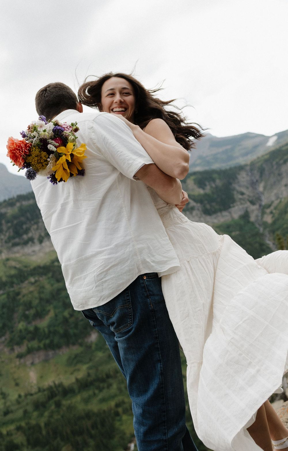 Groom lifts laughing bride holding colorful bouquet with mountain views behind them.