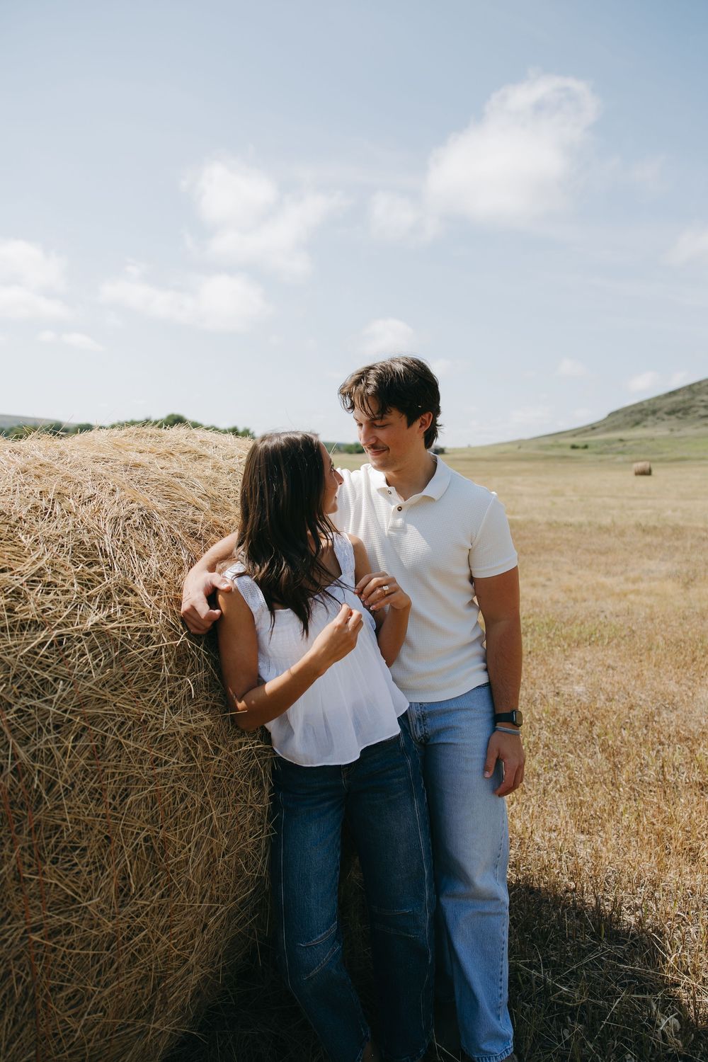 Couple in white tops and jeans leaning against a hay bale in a sunny golden field.