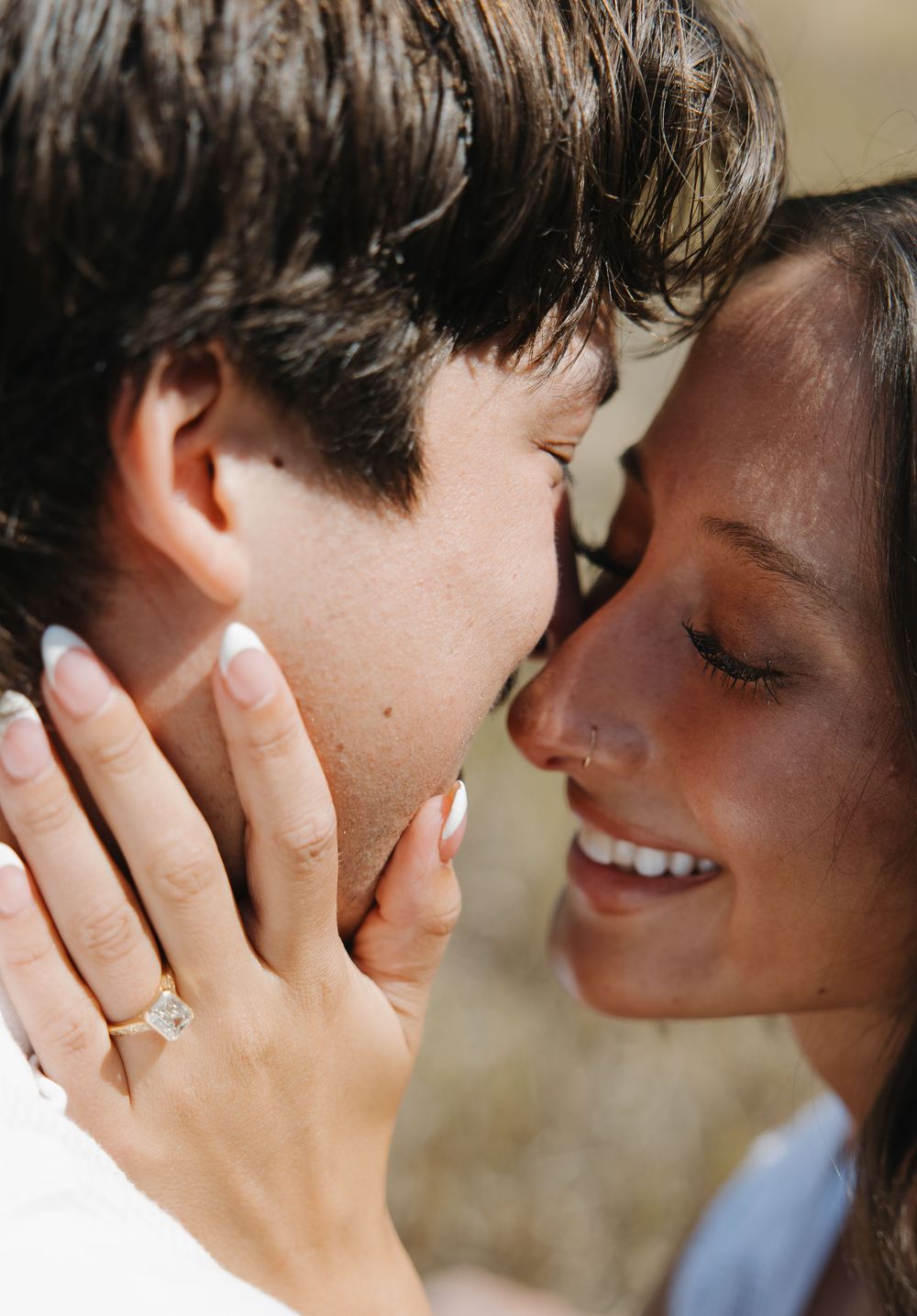 Close-up of smiling couple nearly kissing, woman wearing a gold engagement ring with emerald-cut diamond.