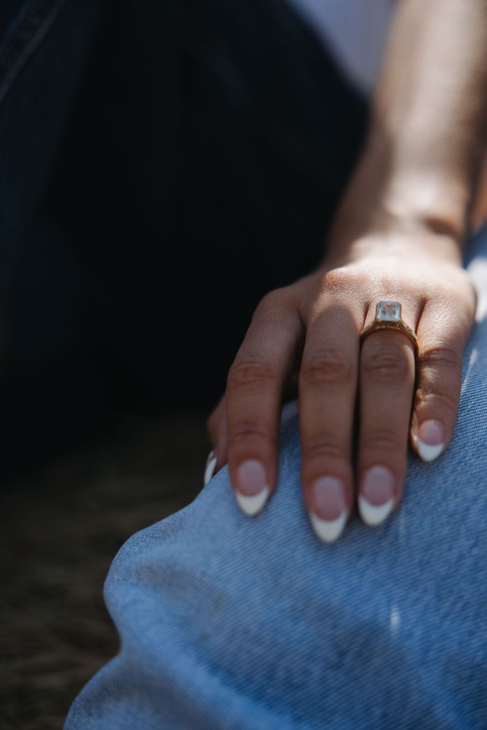 Close-up of womans hand with french manicure and gold emerald-cut engagement ring resting on denim jeans.