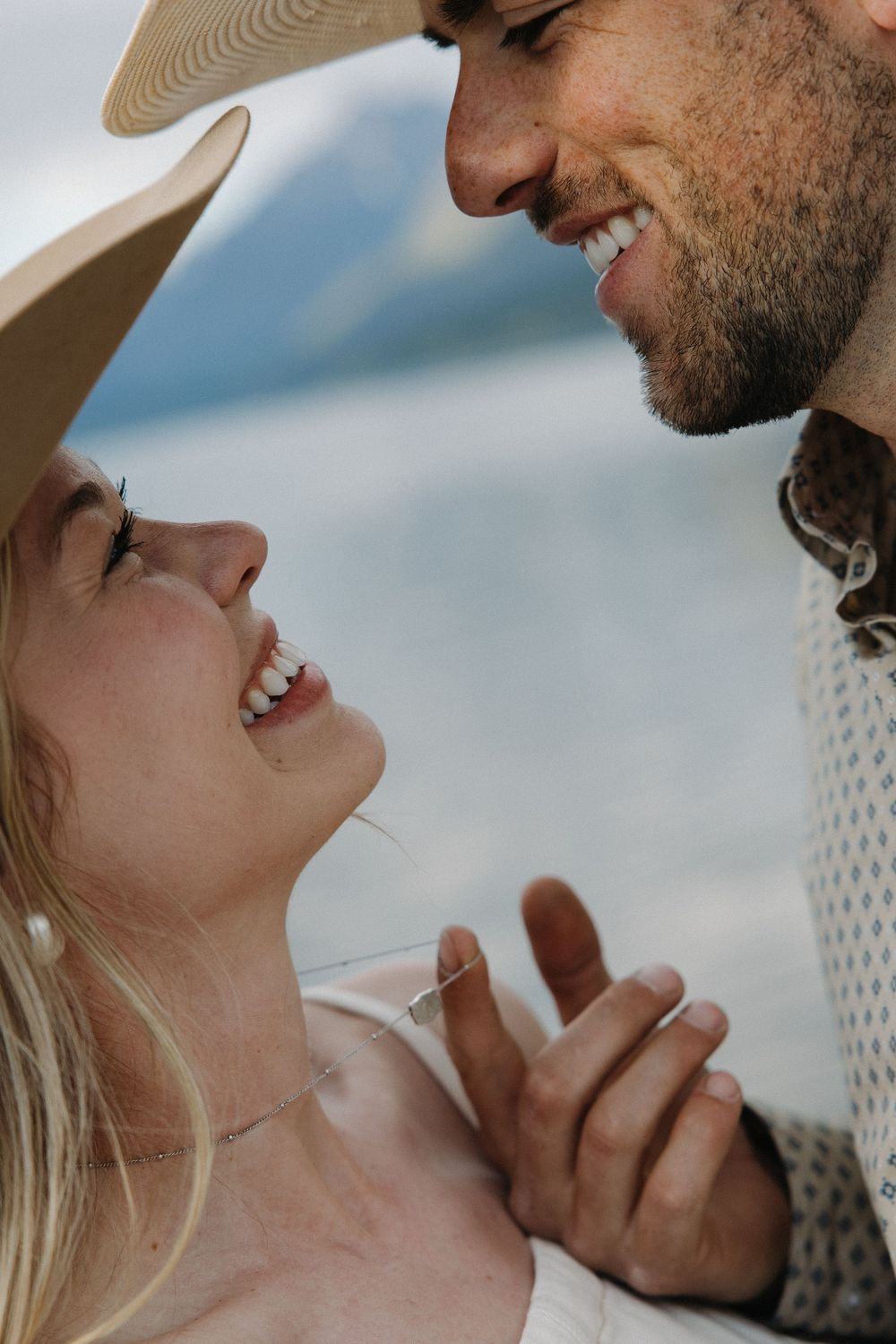 Joyful couple smiling at each other closely, woman wearing a wide-brim hat near a mountain lake.