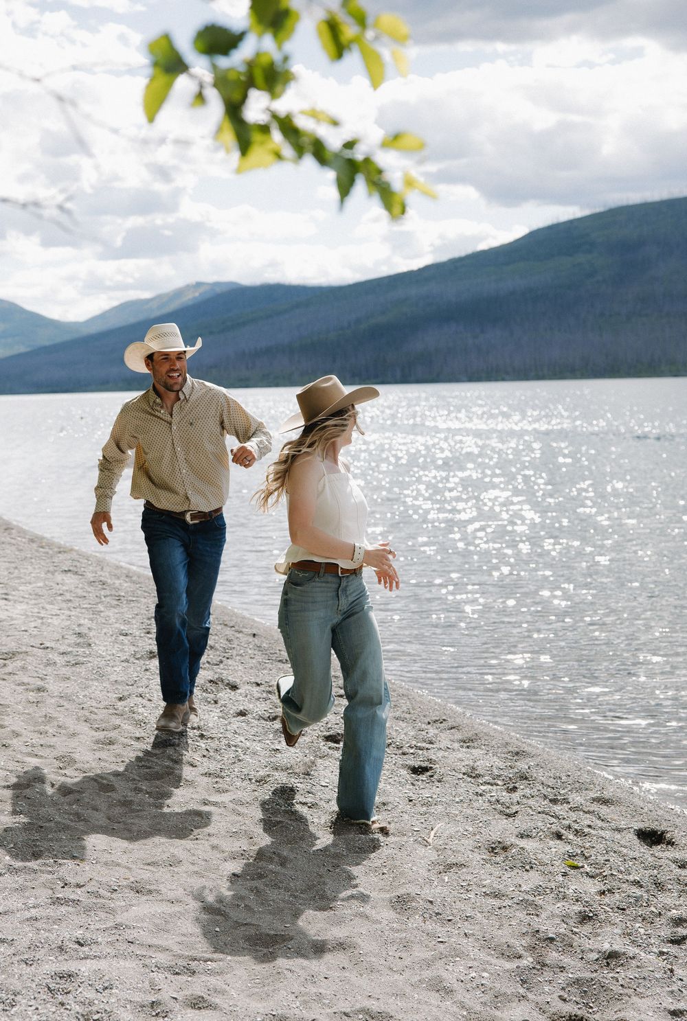 Couple in cowboy hats running playfully along a sandy mountain lakeshore on a cloudy day.