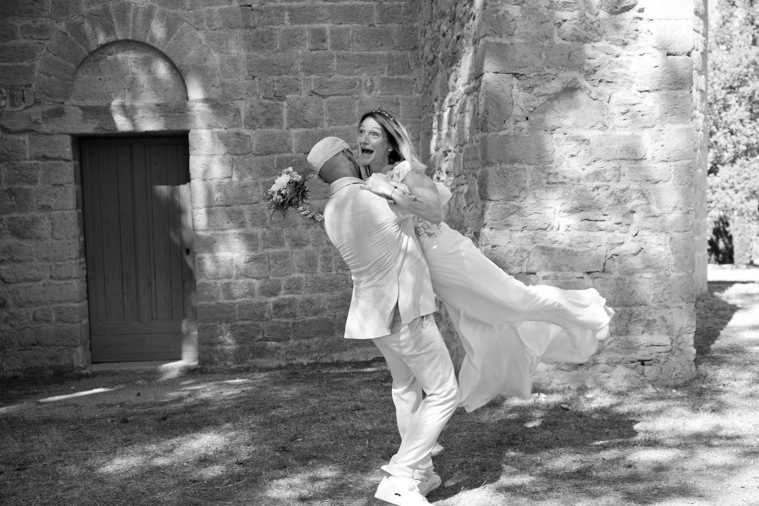Christophe porte Delphine devant une porte ancienne pendant leur séance photo de mariage, en noir et blanc.