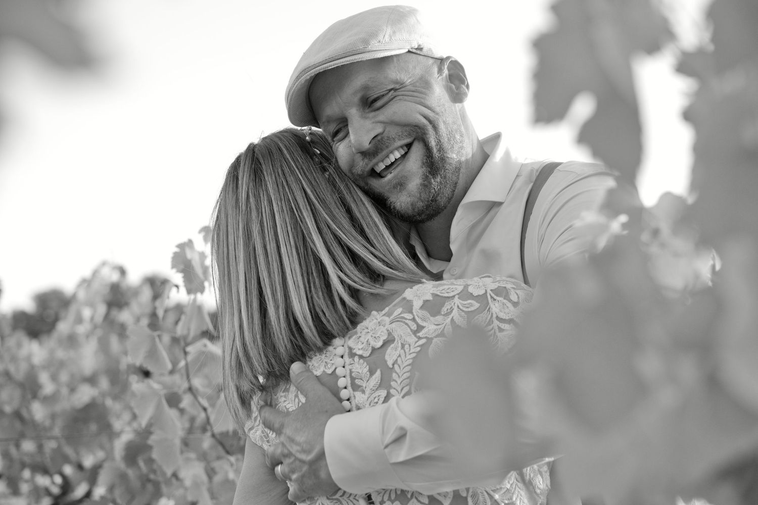 Christophe rit en serrant Delphine dans ses bras au milieu des vignes, en noir et blanc.