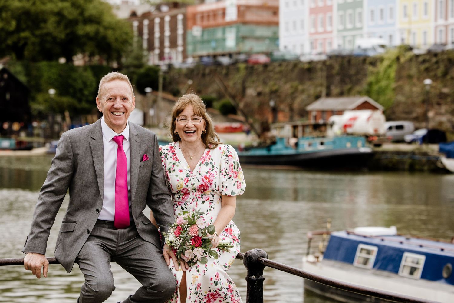 Smiling couple in wedding attire posing by a colorful waterfront harbor with boats and buildings in the background.