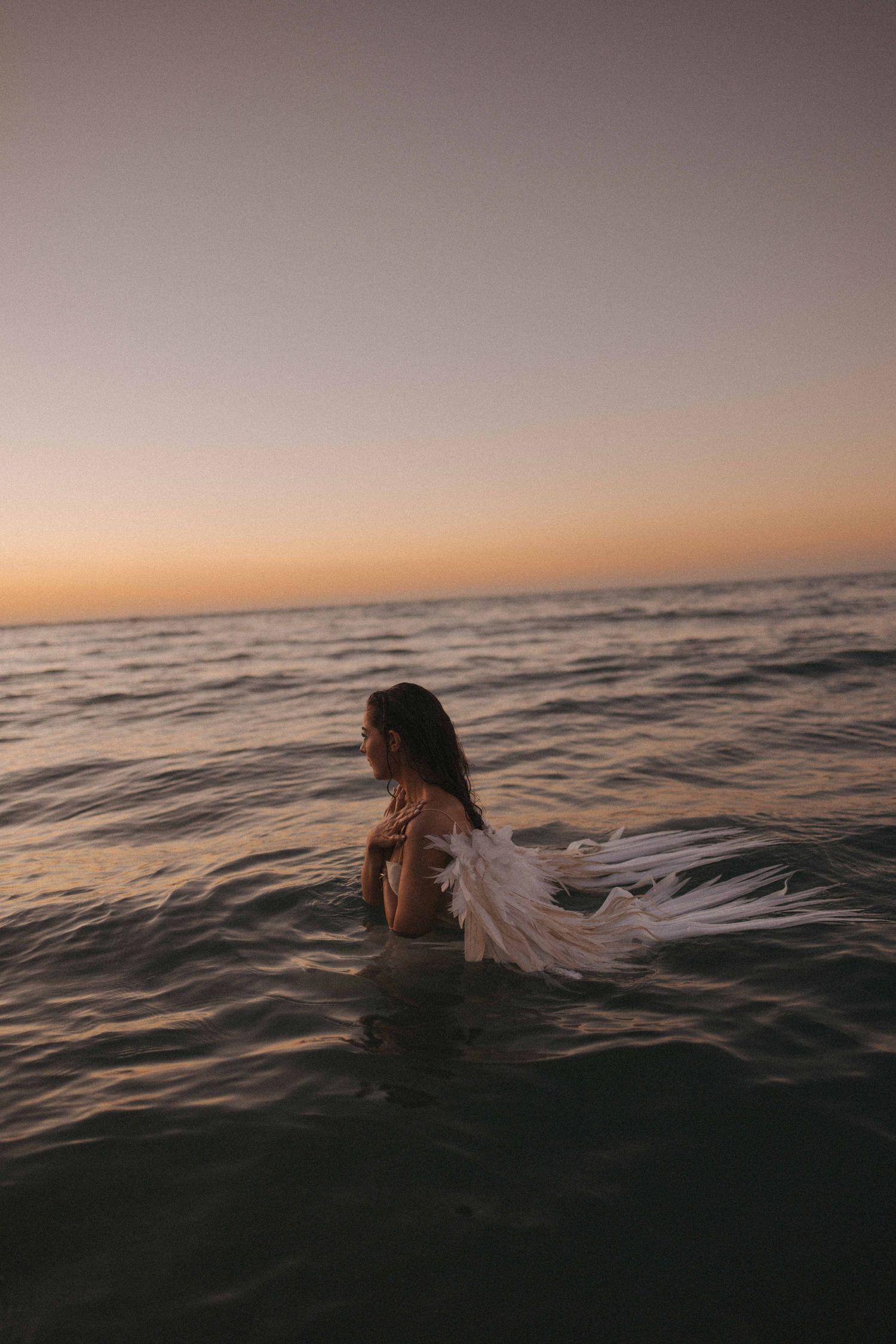 Woman wrapped in a shimmering metallic silver and gold blanket stands on dark rocky shoreline at dusk beneath a pink and purple sky.
