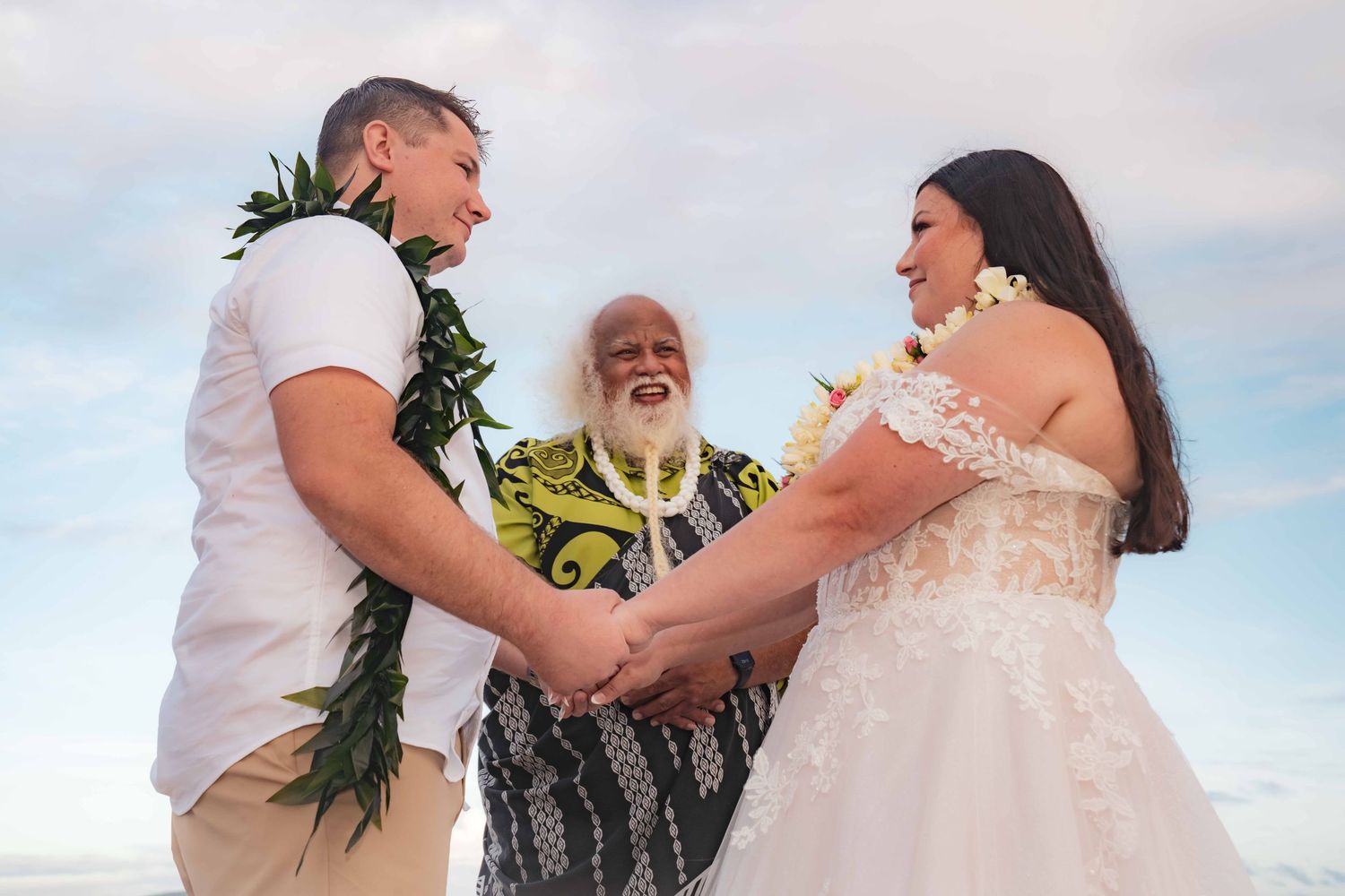 Bride in a white lace gown and groom in a white shirt with a green leaf lei hold hands during a beach wedding ceremony led by an officiant.