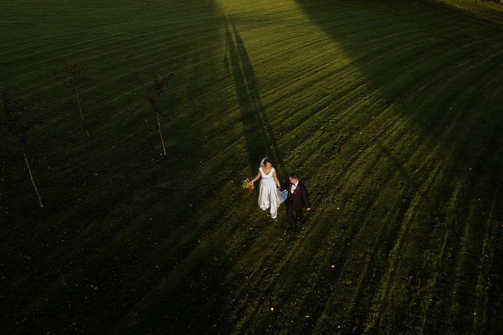 Aerial view of a bride in a white gown and groom walking hand in hand across a green field with long dramatic evening shadows.