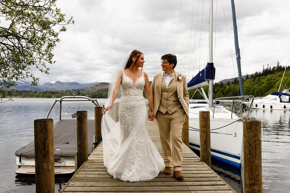 Two newlywed brides in a white lace gown and a tan suit hold hands while walking along a wooden lakeside jetty beside moored sailboats.