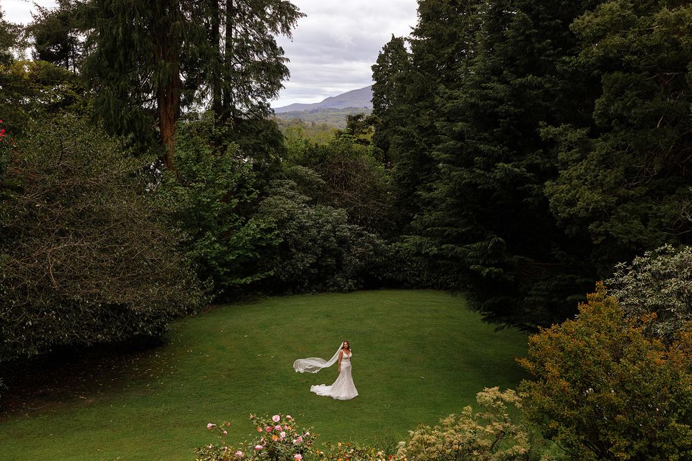 Bride in a white wedding dress with a flowing veil stands on a green lawn surrounded by tall evergreen trees, with misty mountains beyond.