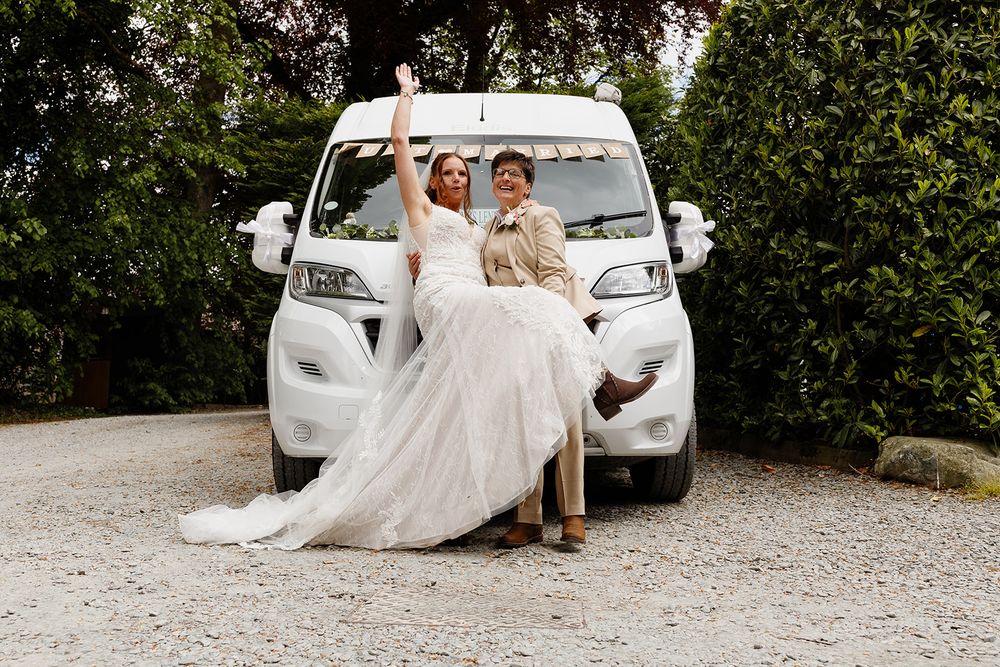 Bride in a flowing white gown raises her arm while seated on the hood of a white van beside a partner in a tan suit, surrounded by greenery.