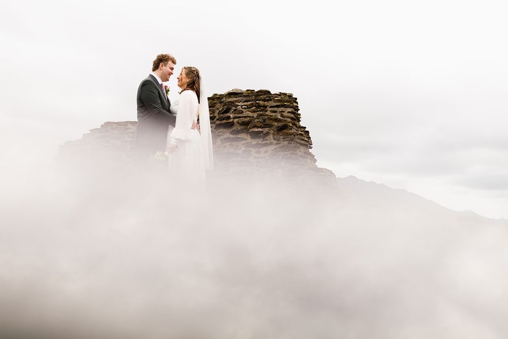 Bride in a white gown and groom in a dark suit stand face to face on a stone ruin rising above thick low clouds.