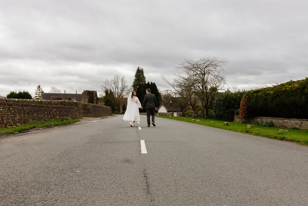 Bride in a white gown and groom in a dark suit walk together down a quiet country road under a cloudy sky.