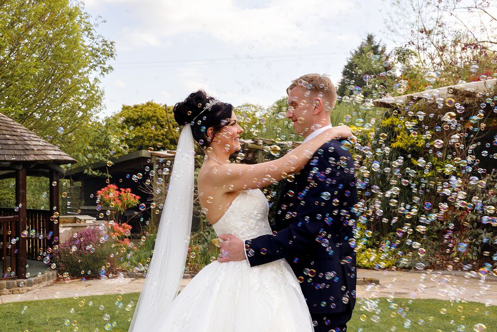 Bride in a white gown and veil embracing a groom in a navy suit in a garden with floral arbors and bubbles floating around them.