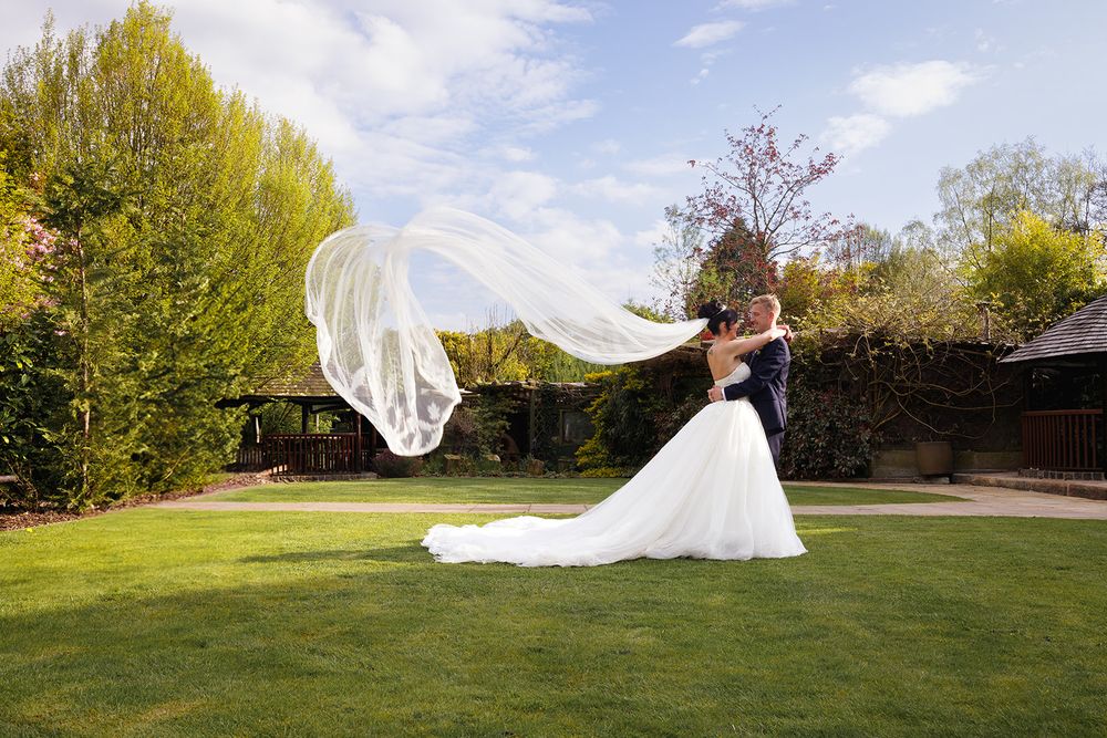 Bride in a flowing white gown embraces her groom on a lush green lawn as her long veil billows in the wind.