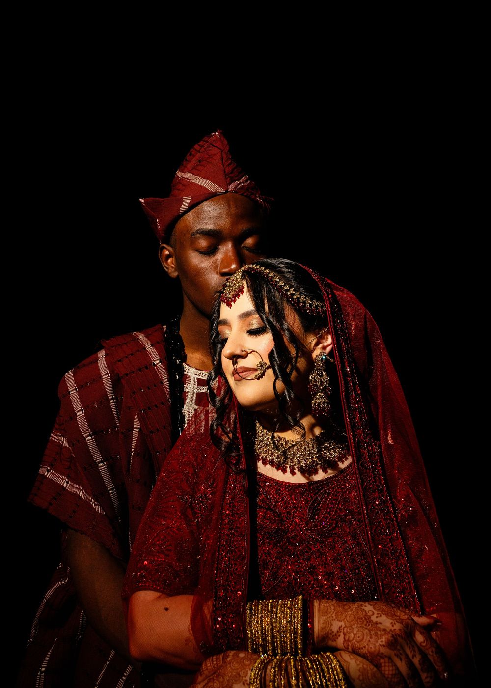 Man in traditional maroon cap and attire gently kisses the forehead of a bride in red embroidered wedding dress with gold jewelry.