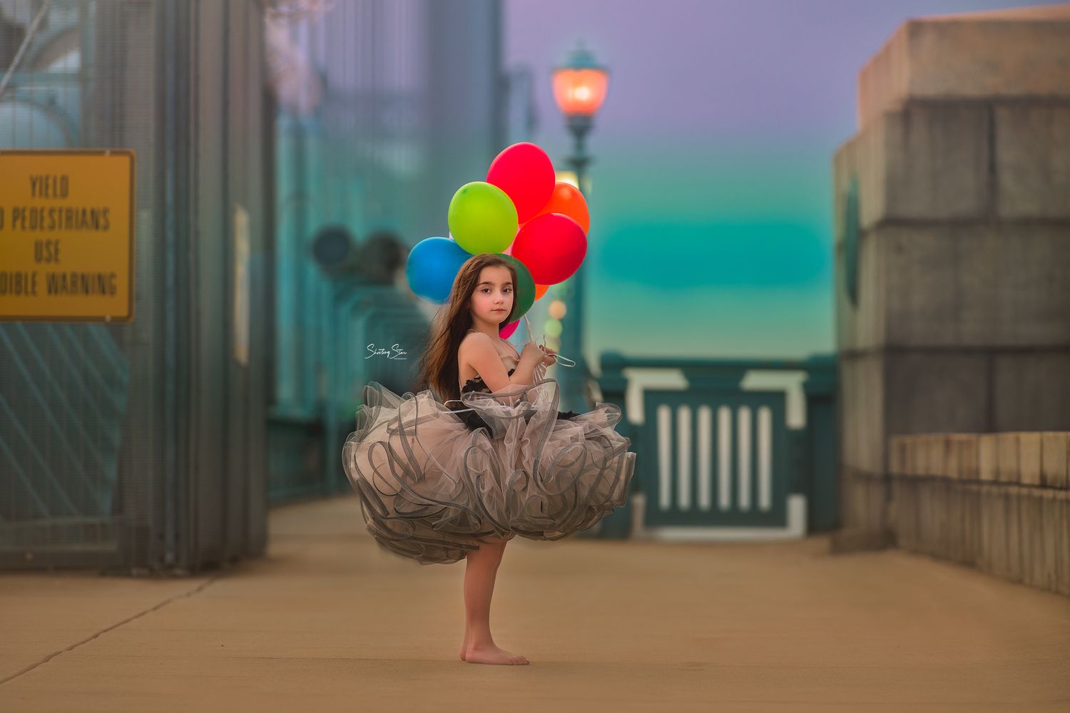 Young girl in a gray tulle tutu skirt holds colorful red, green, and blue balloons while standing barefoot on a city bridge walkway at dusk.