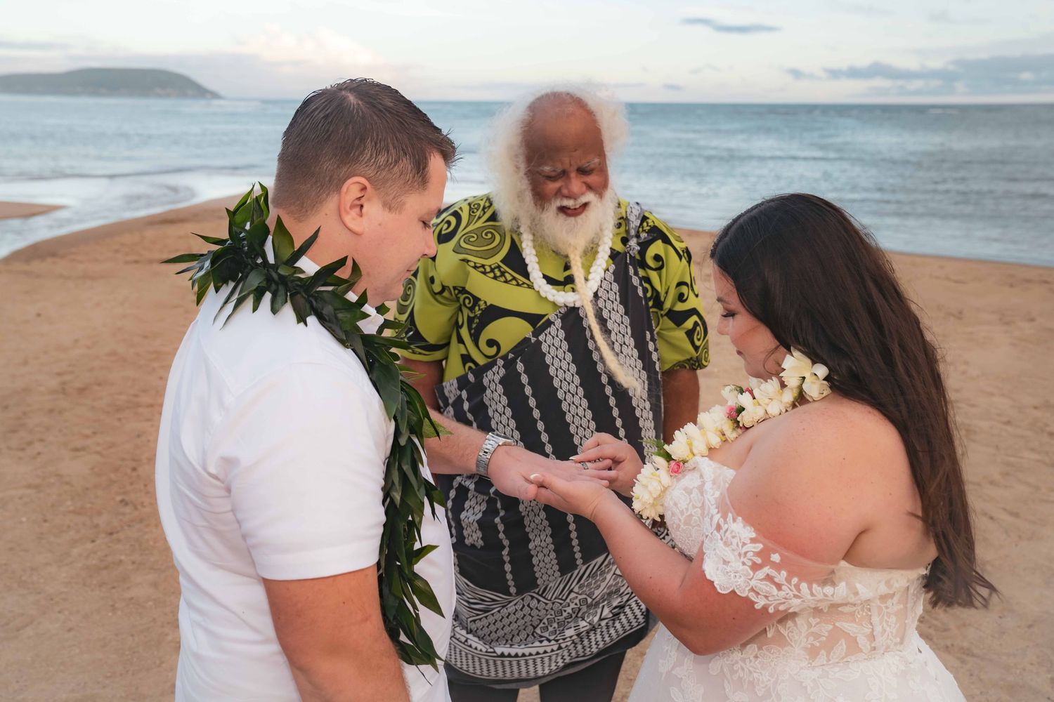 Couple exchanging rings during a beachfront wedding ceremony led by a white-bearded officiant in a floral shirt, with ocean waves behind the