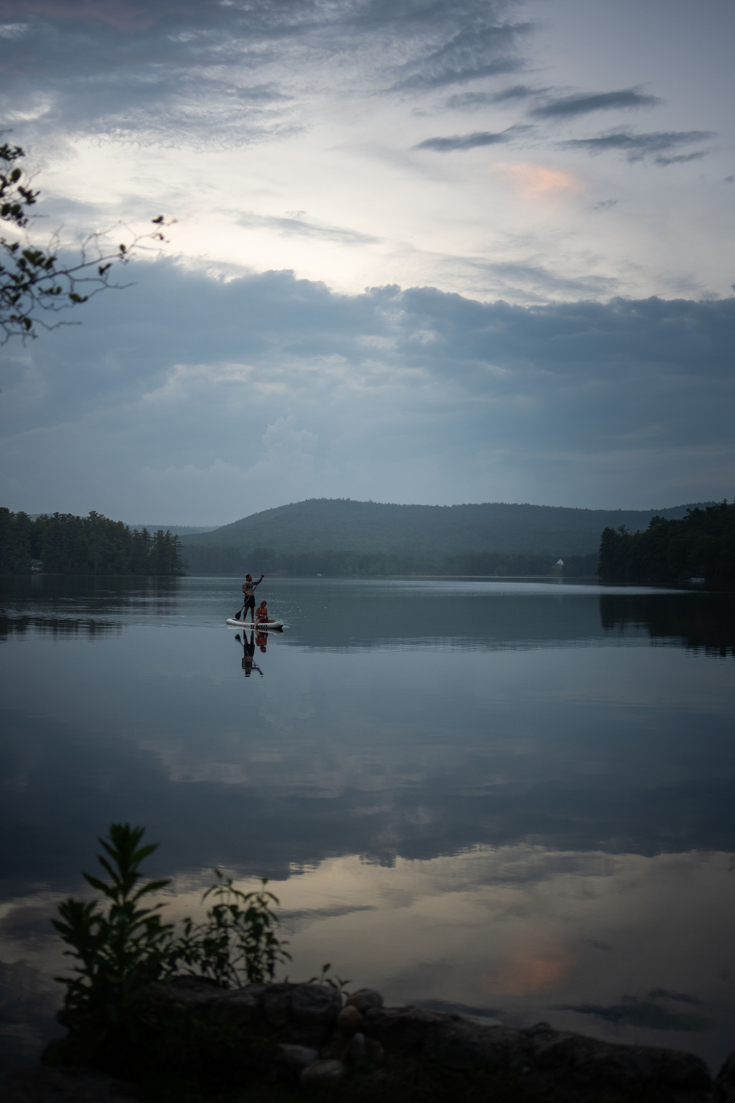 Person paddleboarding on a calm lake at dusk with misty hills and dramatic cloudy sky reflected in still water.