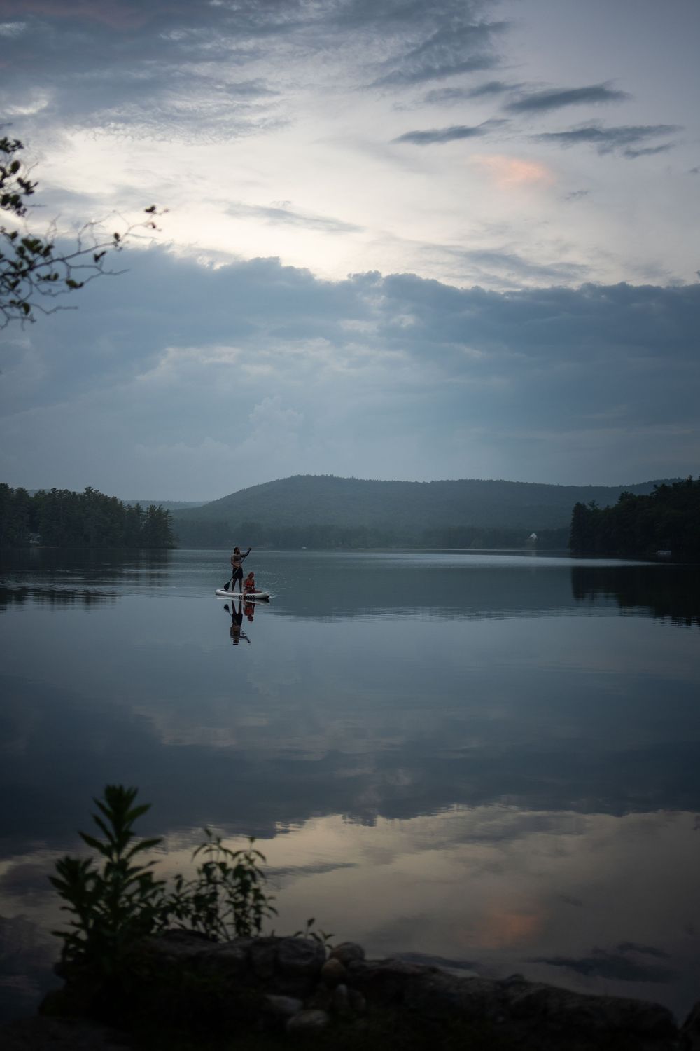 Person paddleboarding on a calm lake at dusk with misty hills and dramatic cloudy sky reflected in still water.