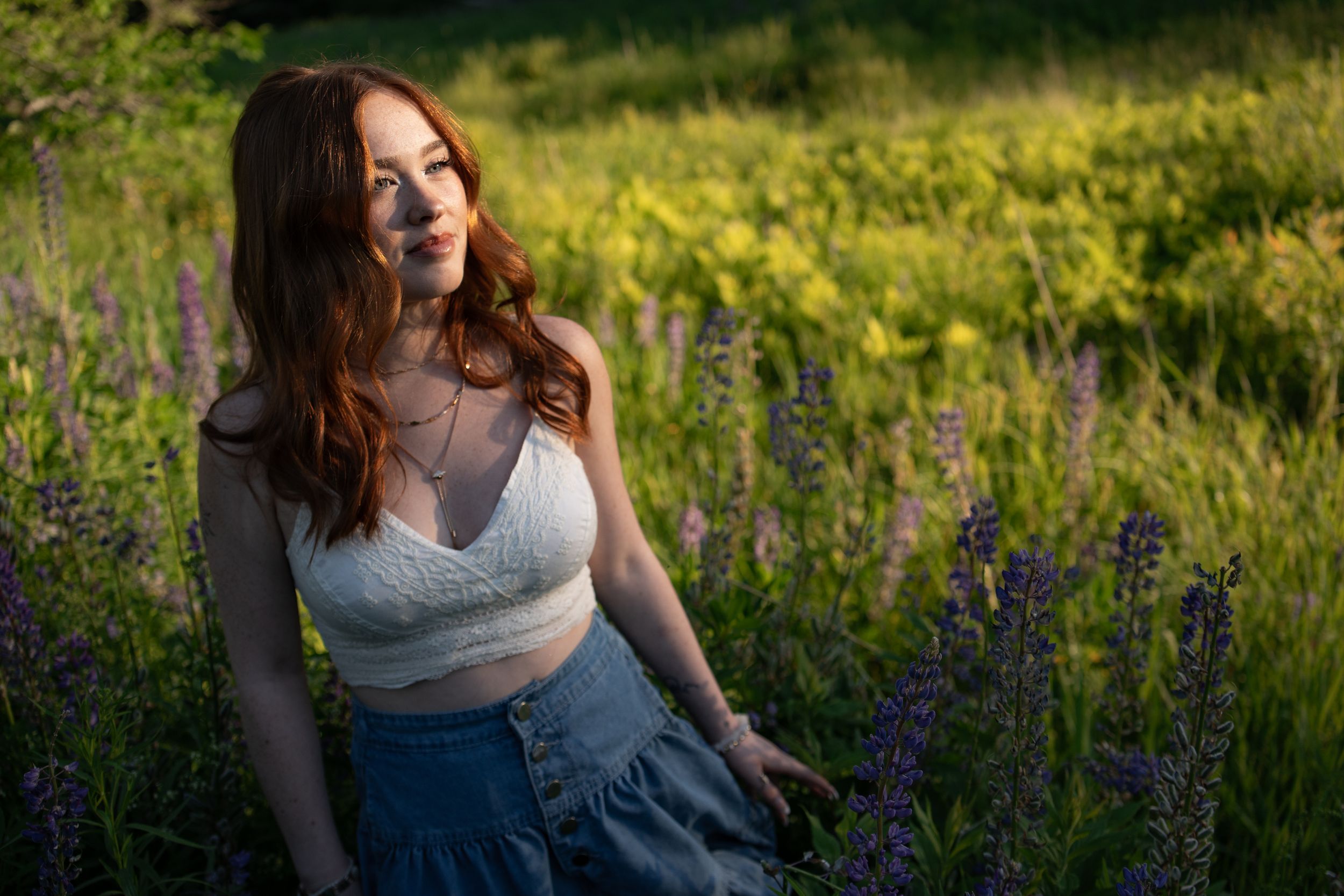 A young woman with red hair sits among purple wildflowers in a sunlit meadow wearing a white crop top and blue skirt.