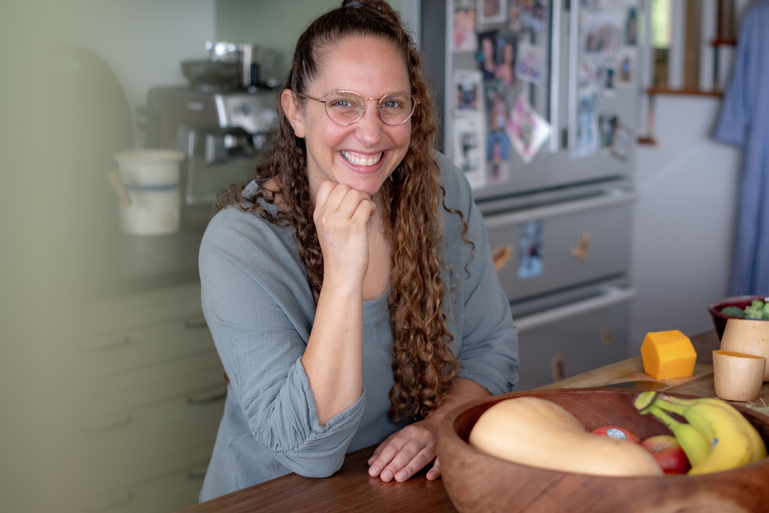Smiling woman with curly hair and glasses leaning on a wooden counter with fresh fruits in a cozy kitchen.