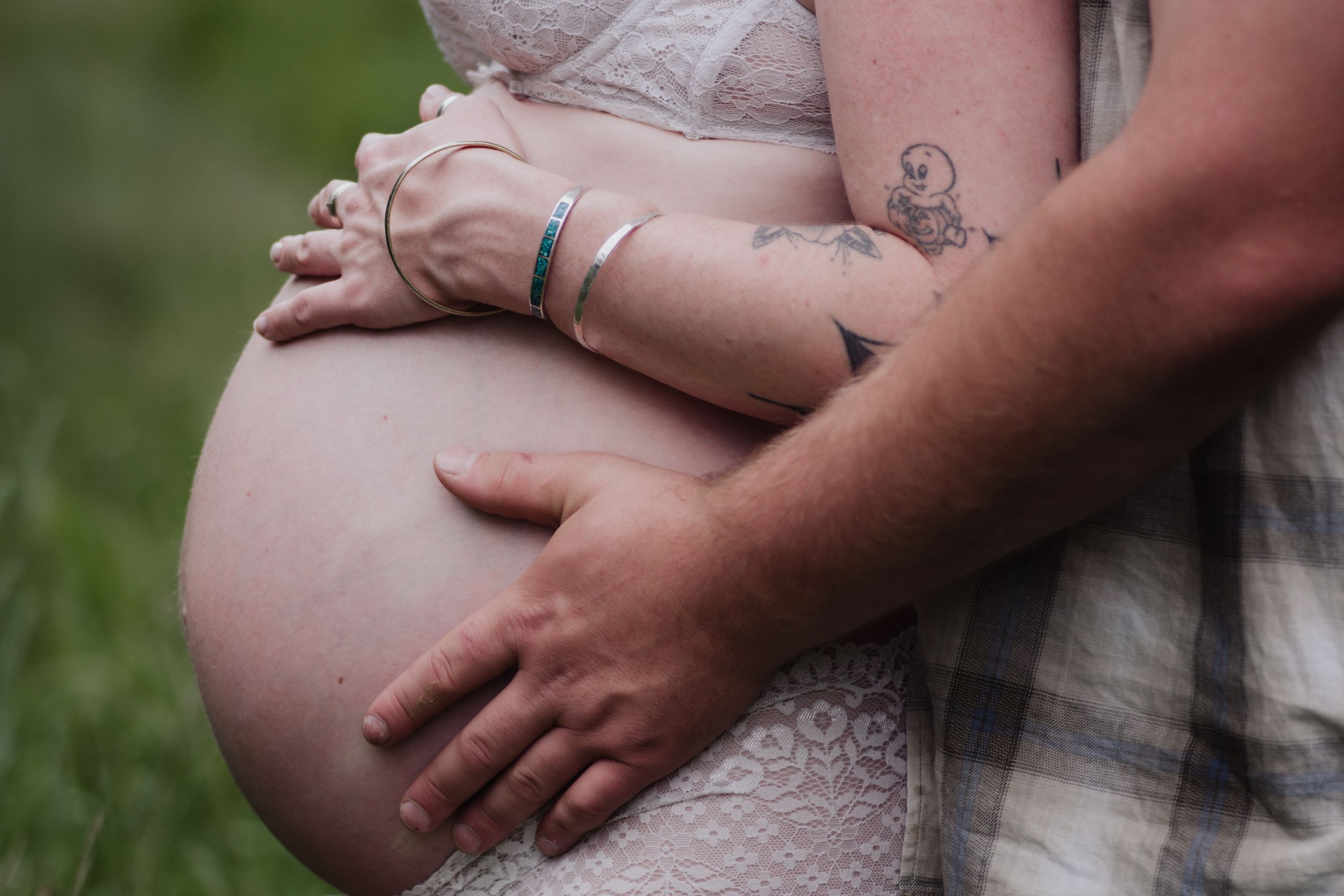 Pregnant woman in lace dress with partner's hand on baby bump outdoors.