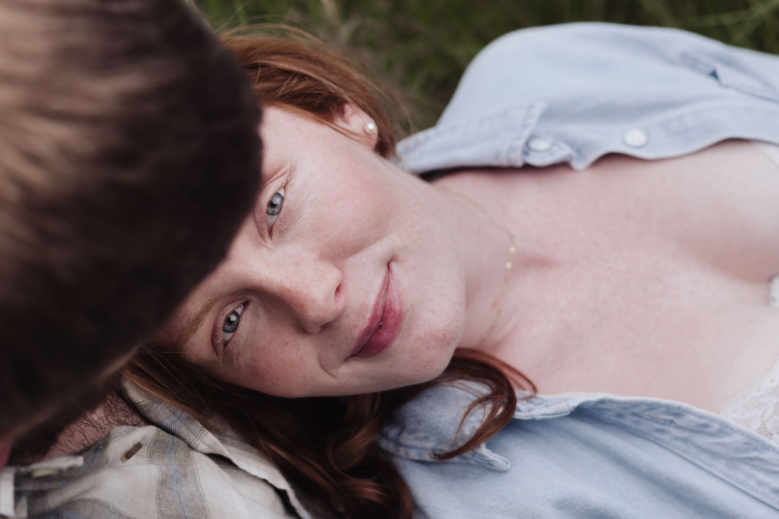 A young redhead woman with blue eyes and freckles lying in grass, looking up at the camera.