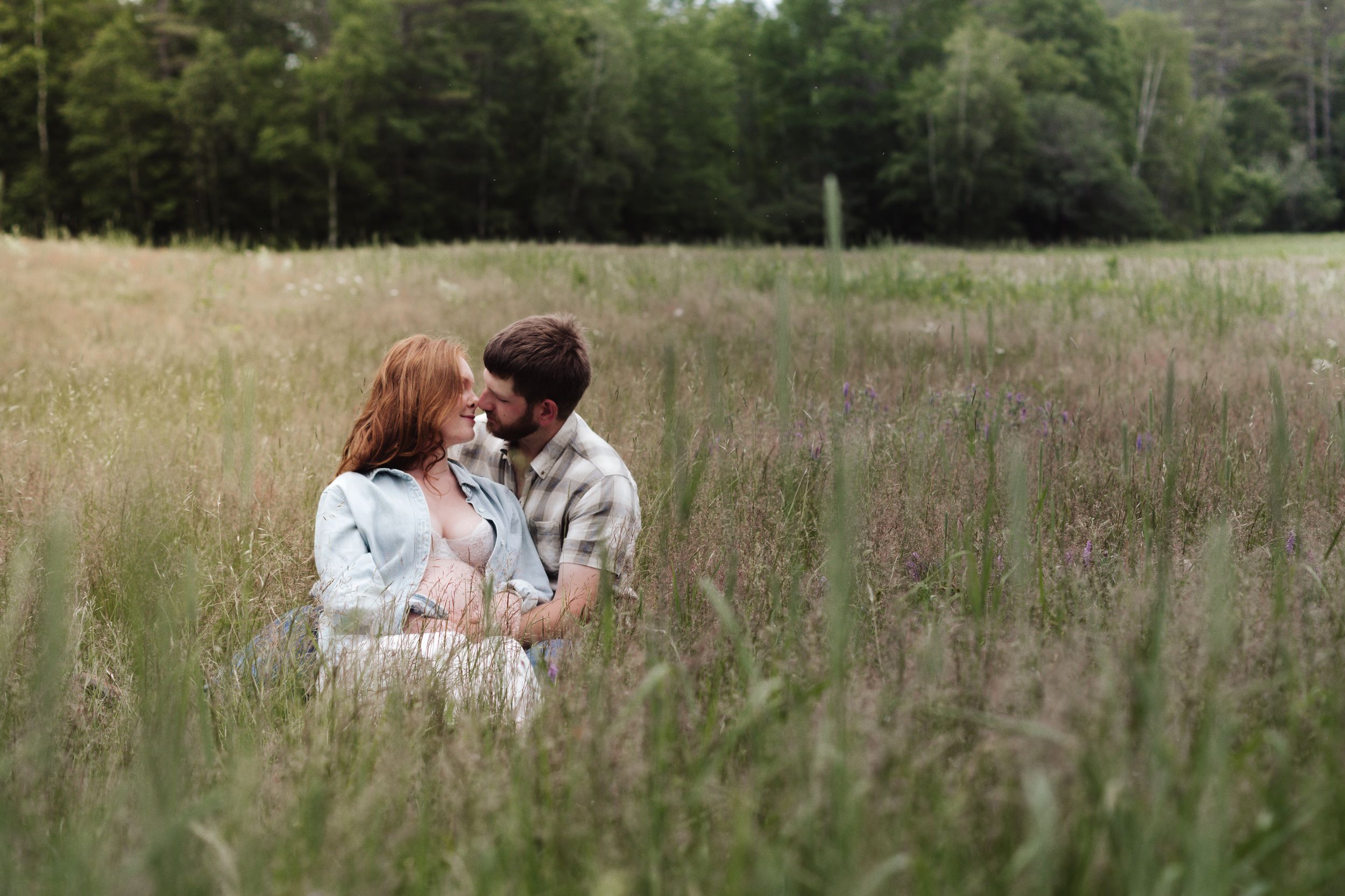 Couple sitting together in a lush green meadow, sharing a romantic moment surrounded by tall grass and trees.