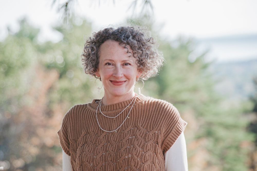 Smiling woman with curly gray hair wearing a brown knit vest and layered necklaces outdoors.