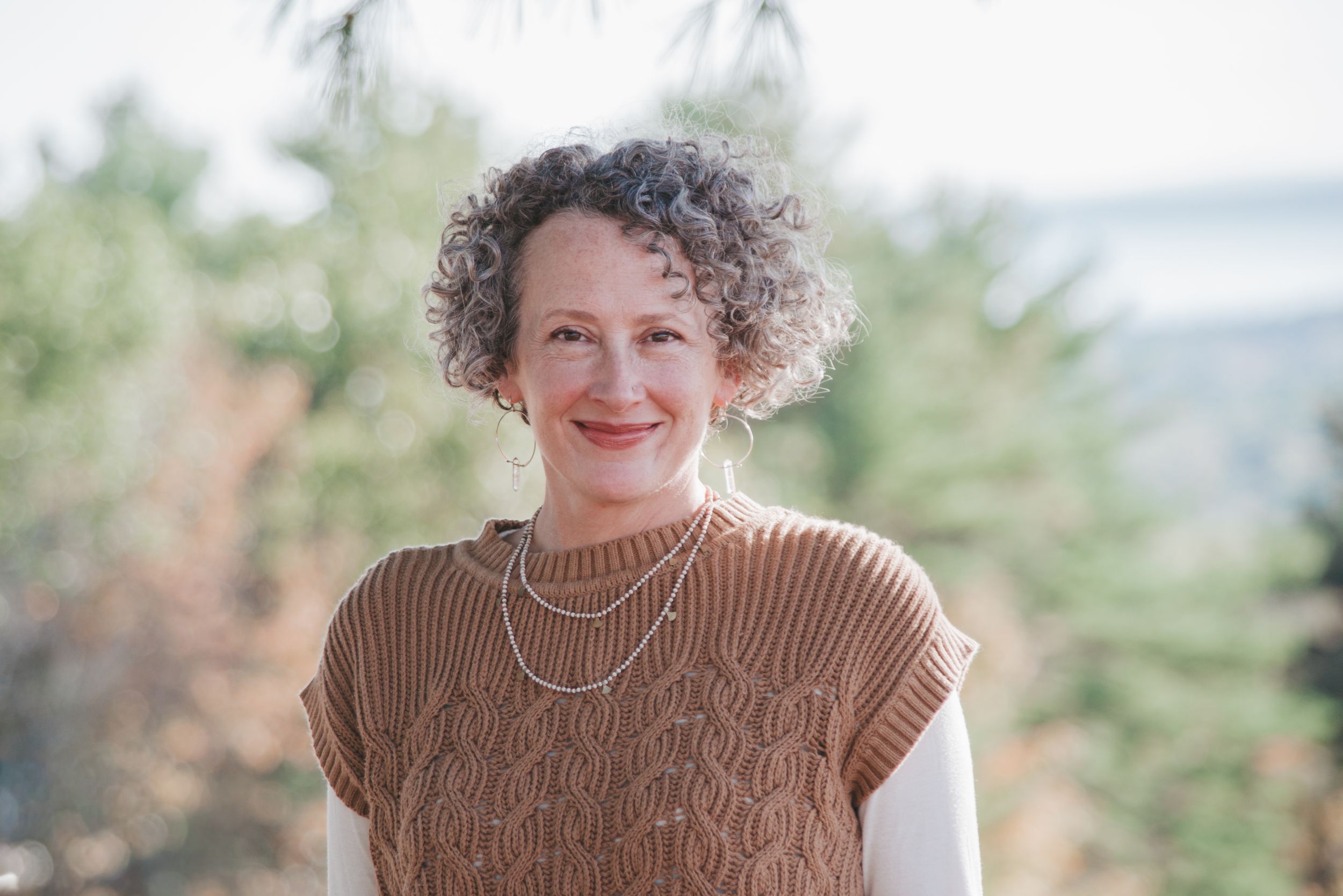 Smiling woman with curly gray hair wearing a brown knit vest and layered necklaces outdoors.