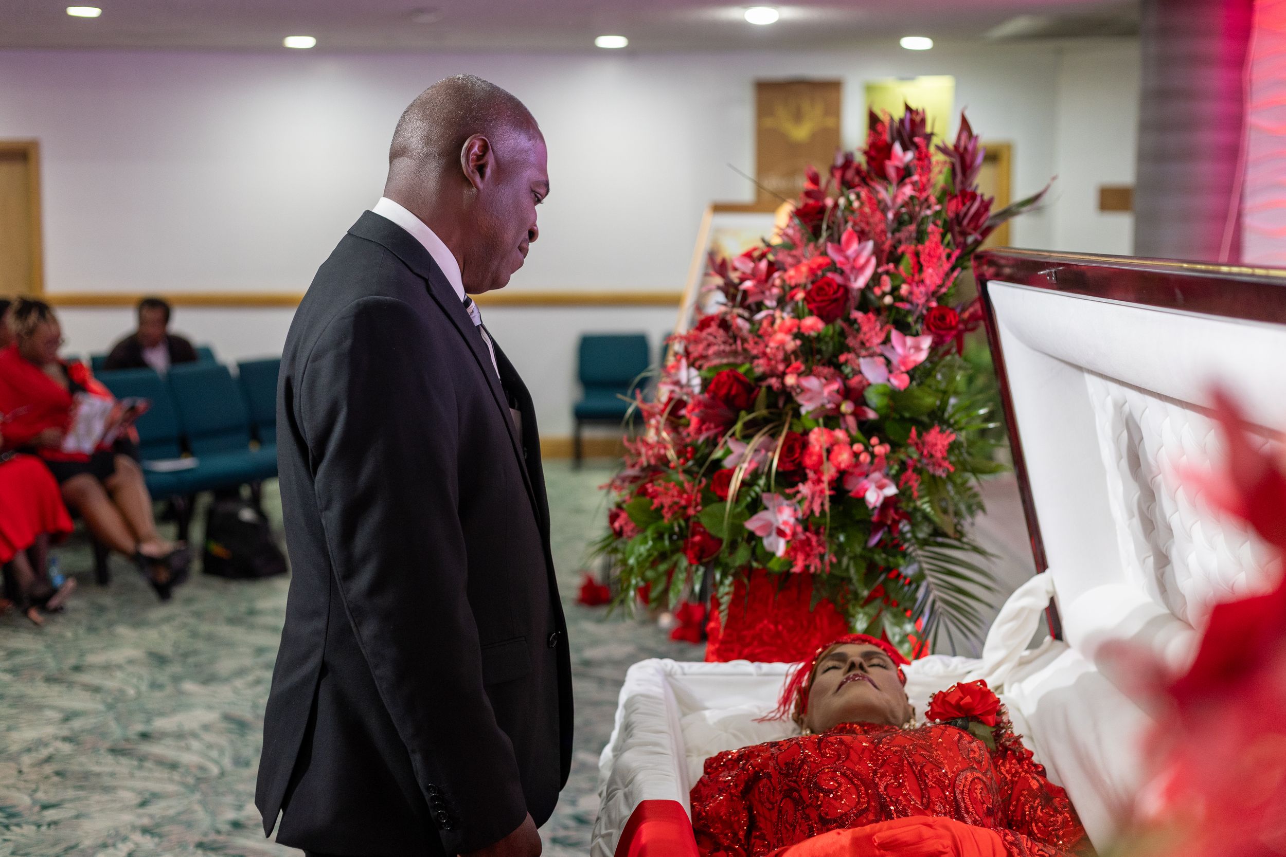 Funeral service setup with red floral arrangements and black casket in a formal indoor setting.